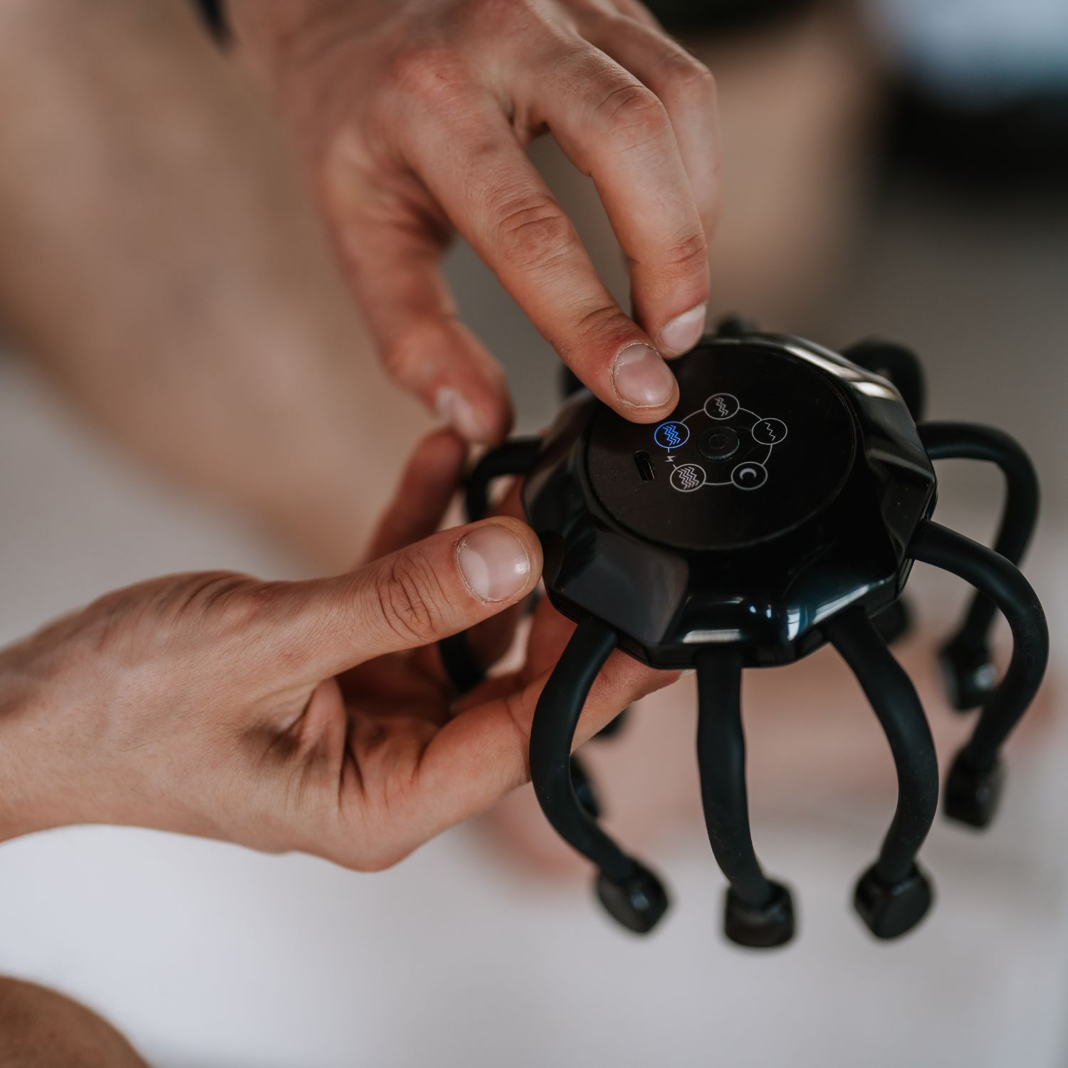 Hände halten ein schwarzes Kopfmassagegerät. Eine Hand drückt einen Knopf auf dem Gerät. Das Massagegerät hat flexible Beine und ein zentrales Bedienfeld.