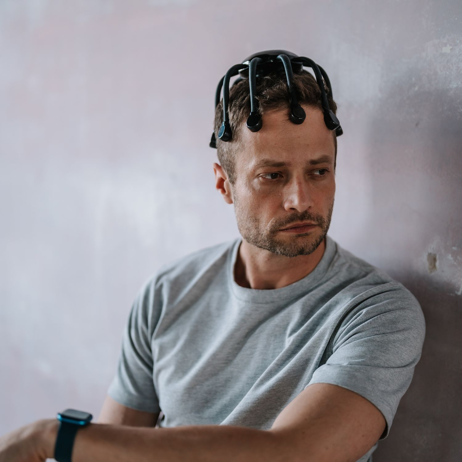 Ein Mann mit schwarzer EEG-Kappe, grauem T-Shirt und Uhr lehnt an einer rosa-grauen Wand und blickt zur Seite.