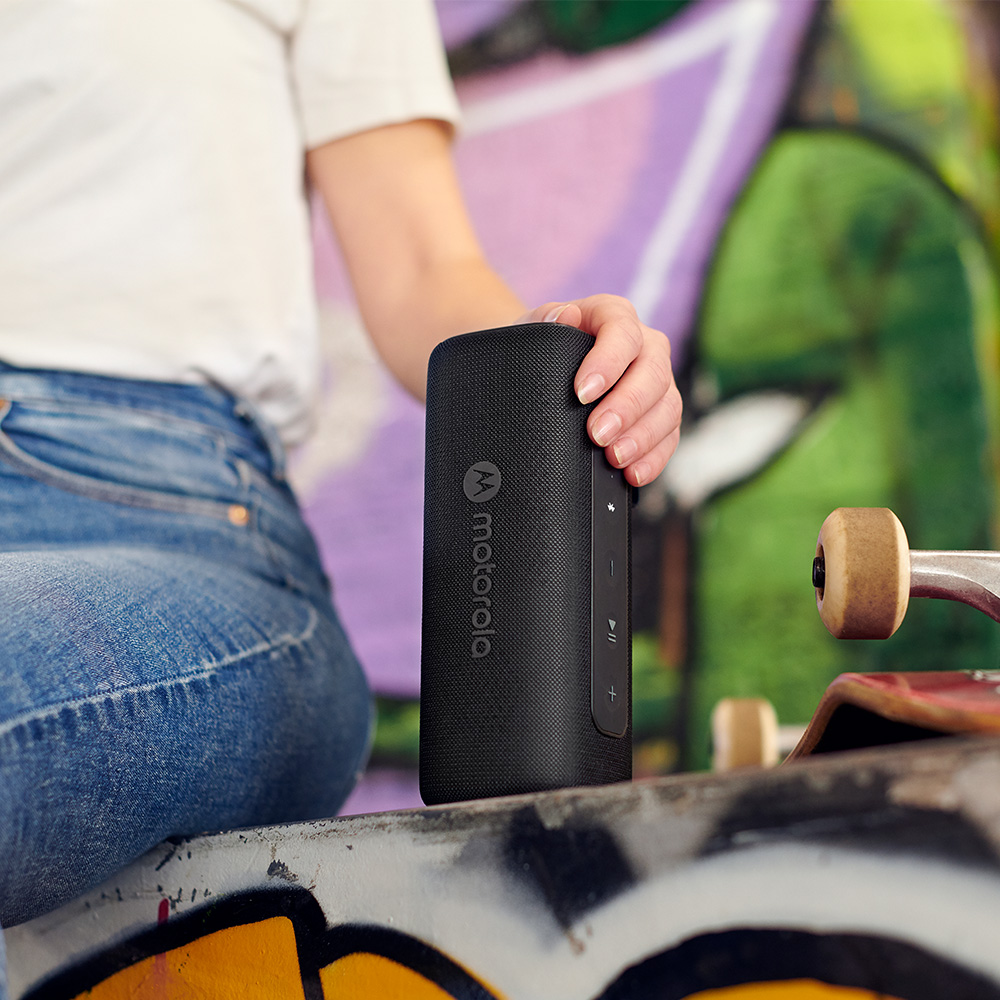 Eine Person hält einen schwarzen Motorola-Lautsprecher. Der Lautsprecher liegt auf einem Skateboard. Im Hintergrund ist eine bunte Graffiti-Wand zu sehen.
