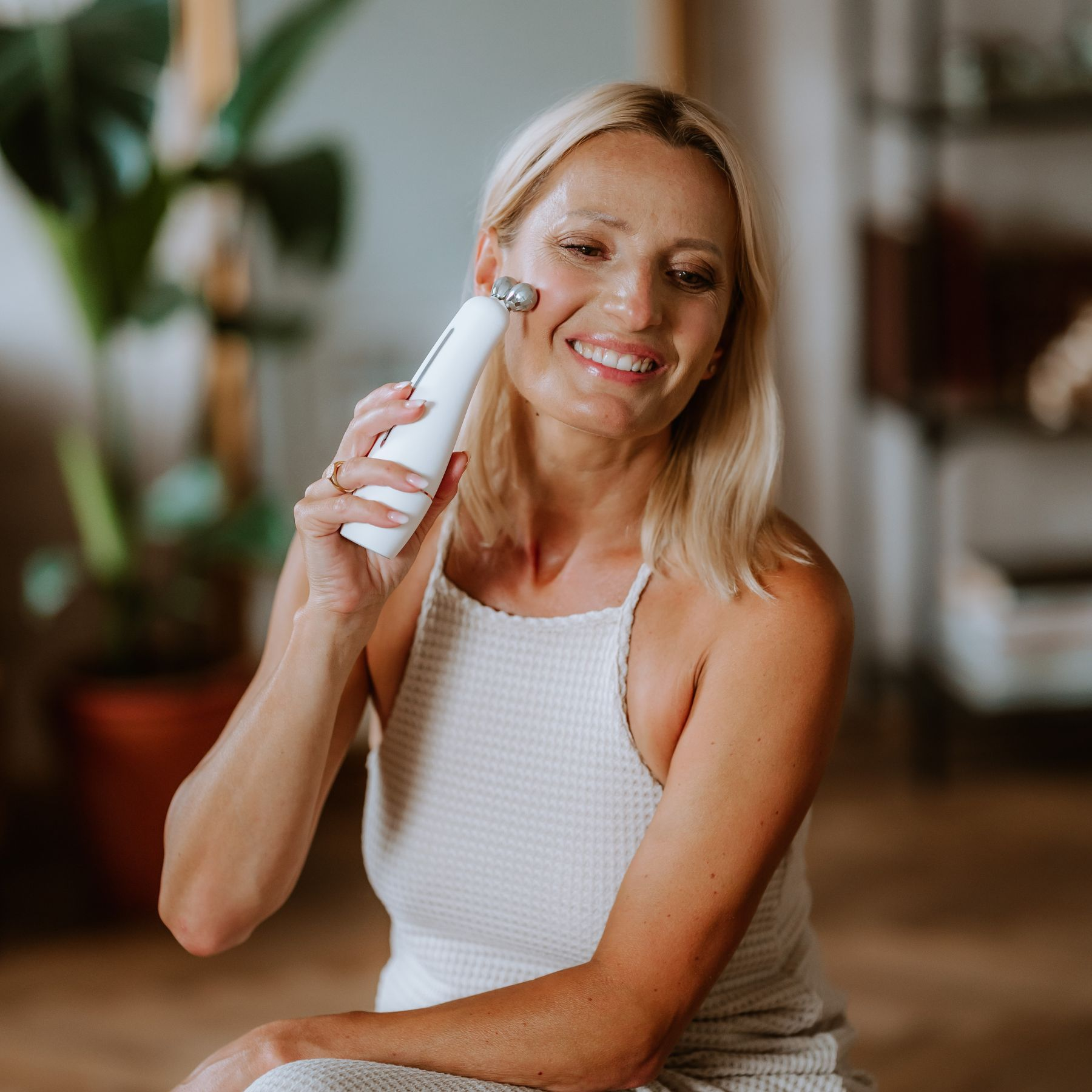 Eine lächelnde Frau benutzt ein weißes Gesichtsmassagegerät. Sie hat blonde Haare und trägt ein weißes Oberteil. Im Hintergrund ist eine Pflanze.