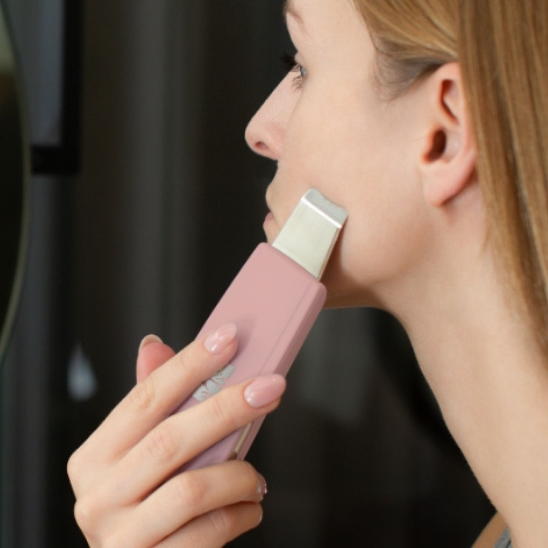 Kobieta używa różowego narzędzia do pielęgnacji skóry na twarzy. Narzędzie ma srebrną końcówkę. Trzyma je z wypielęgnowanymi paznokciami.