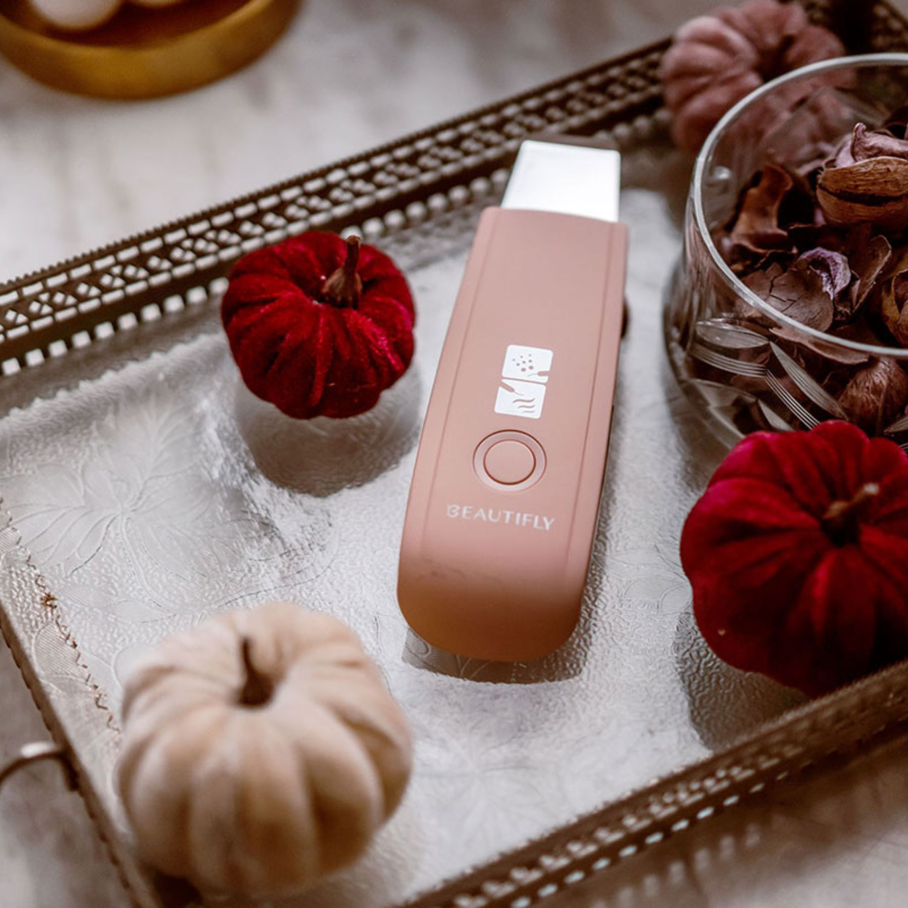 Różowe urządzenie kosmetyczne na tacy z aksamitnymi dyniami i szklaną miską z suszonymi roślinami.