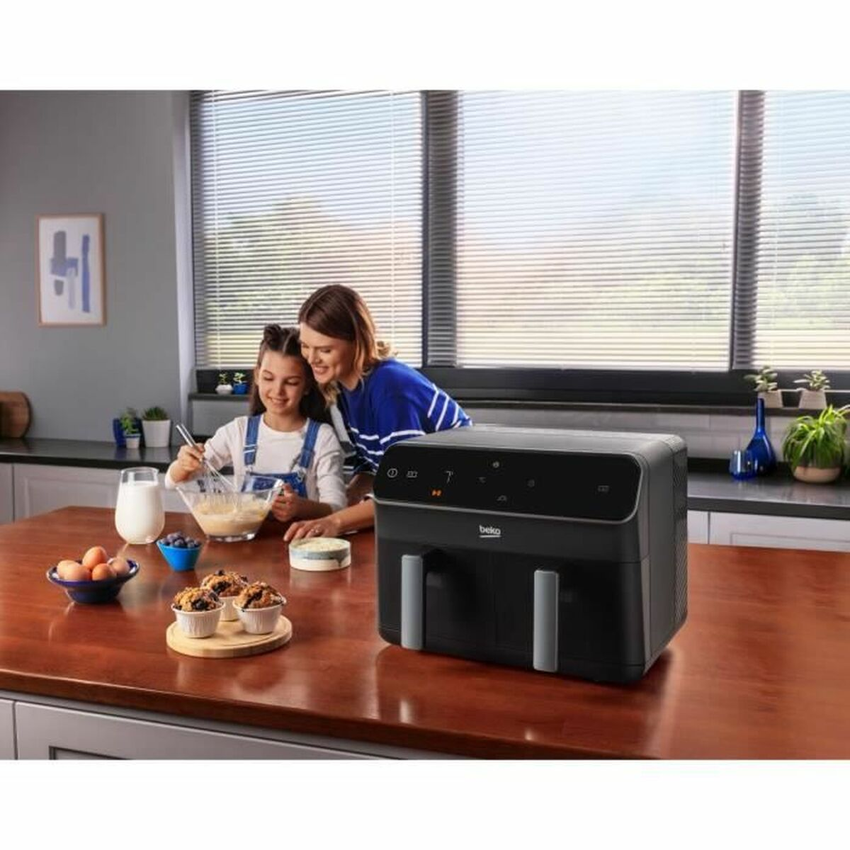 A woman and girl are baking. An air fryer sits on the table. Ingredients include eggs, milk, and muffins.