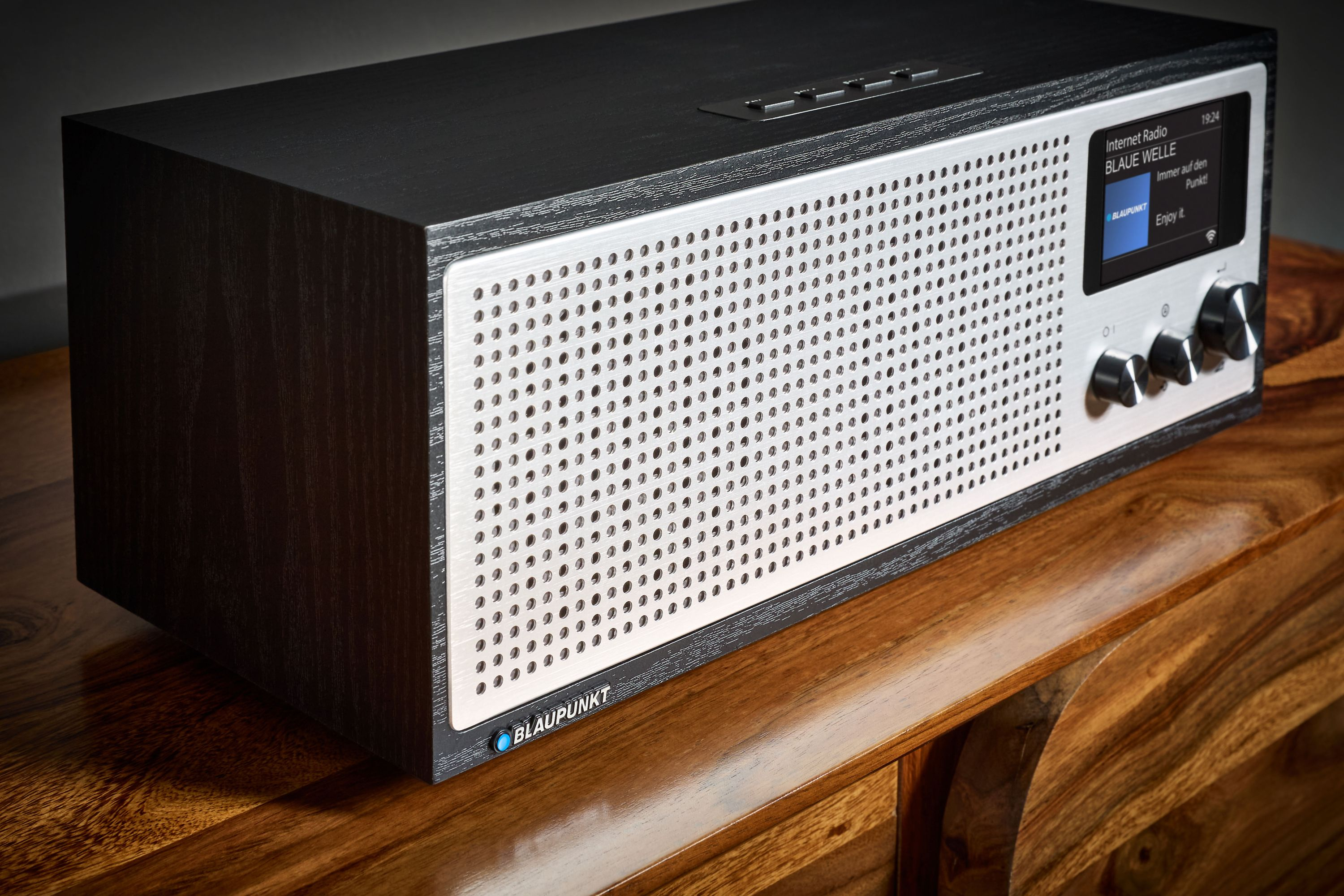 Ein schwarz-silbernes Radio auf einem Holztisch. Das Radio hat ein Display mit Radiosenderinformationen.