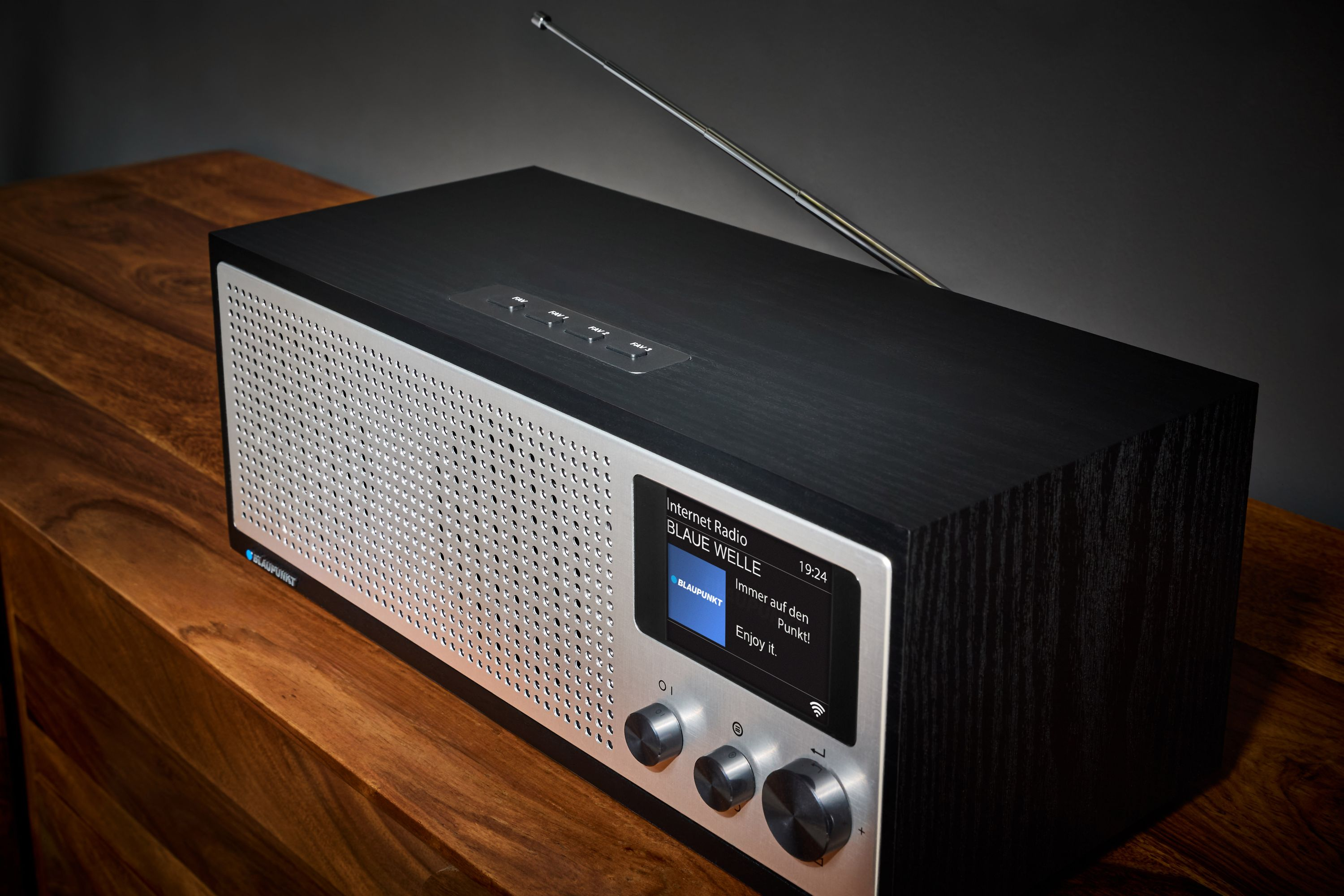 Ein schwarzes und silbernes Radio auf einer Holzoberfläche. Ein Bildschirm zeigt Informationen an. Eine Antenne ist ausgefahren.