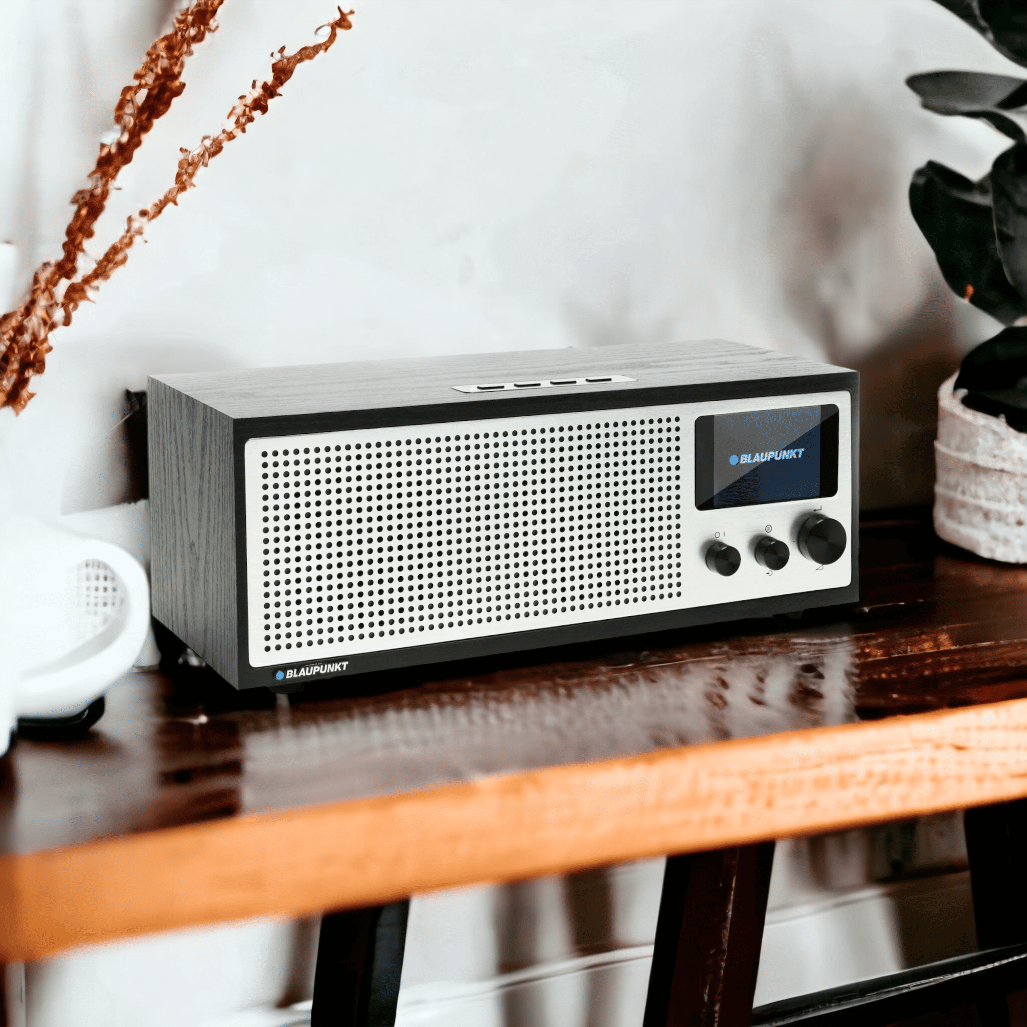 Ein Blaupunkt-Radio auf einem Holztisch. Es hat ein Display und Knöpfe. Pflanzen und dekorative Elemente sind im Hintergrund.