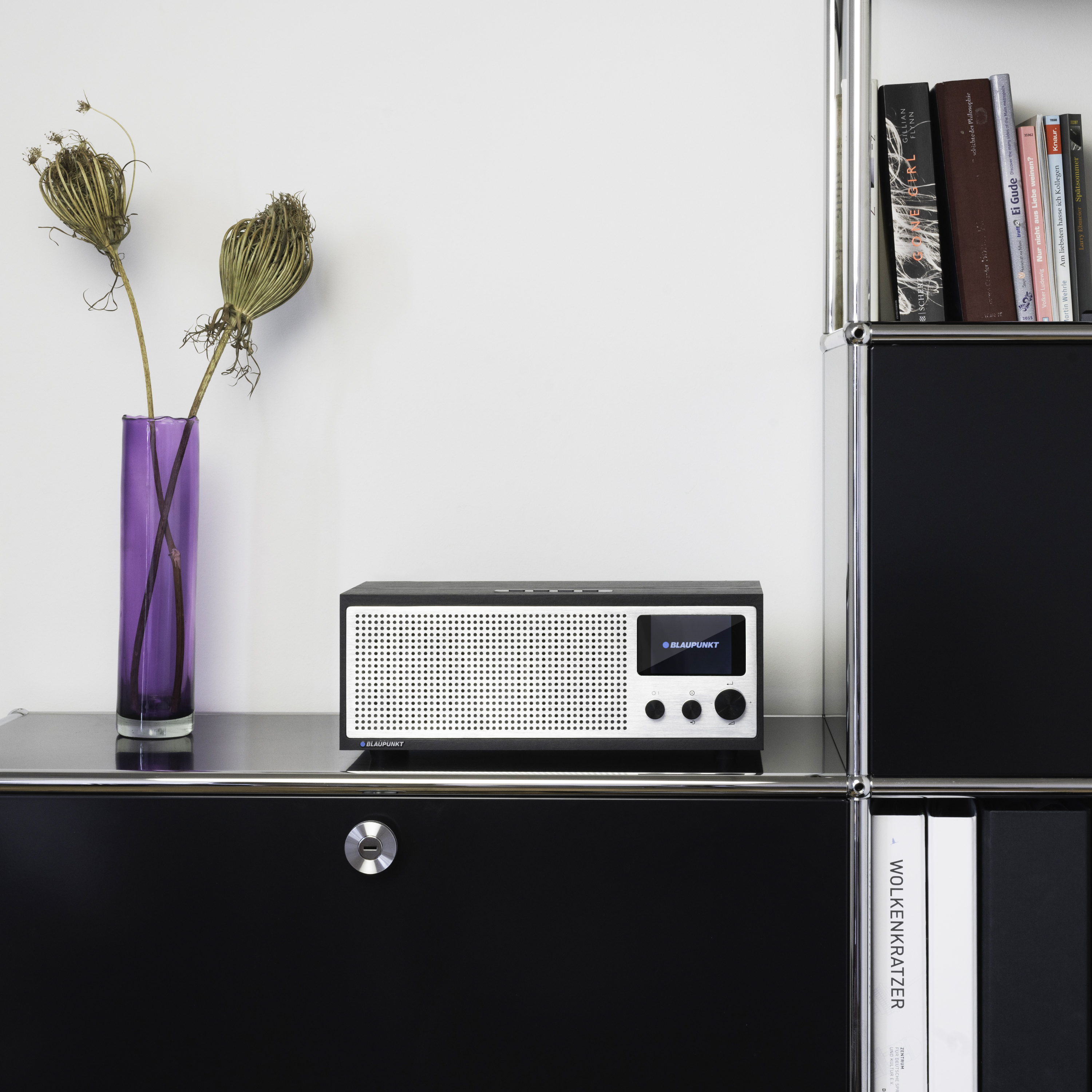 Ein schwarzes Radio mit weißem Lautsprecher neben einer lila Vase mit getrockneten Blumen. Bücher auf Regalen.