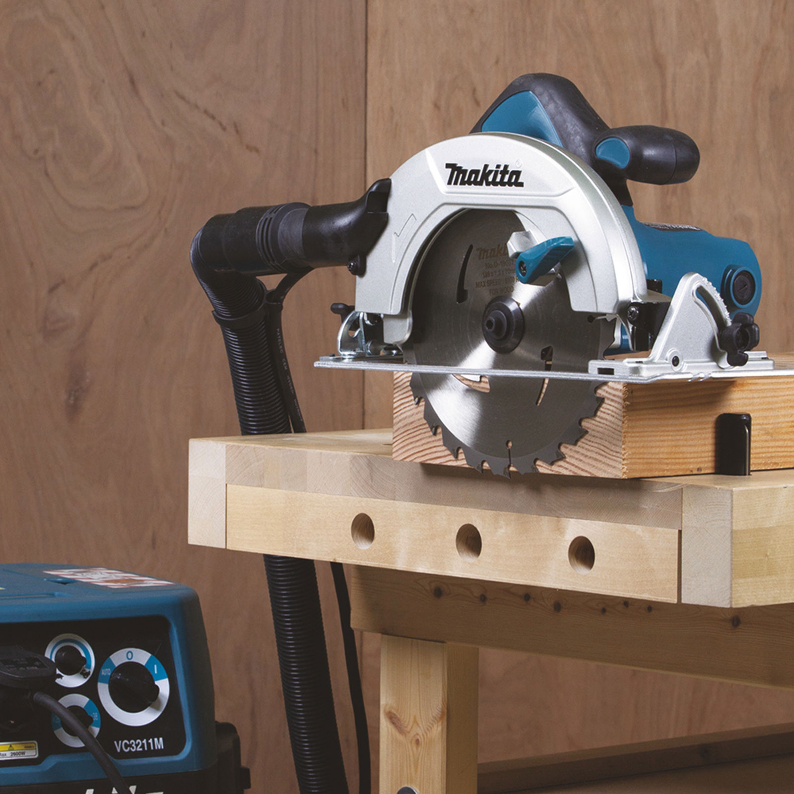 A Makita circular saw on a wooden workbench, with a vacuum cleaner on the left. The saw is cutting a wooden block.