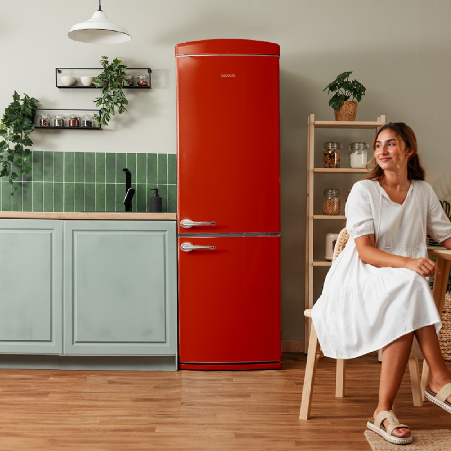 Una cucina con frigorifero rosso, donna in abito bianco, scaffali in legno, piante e paraschizzi verde. Pavimento in legno e illuminazione.