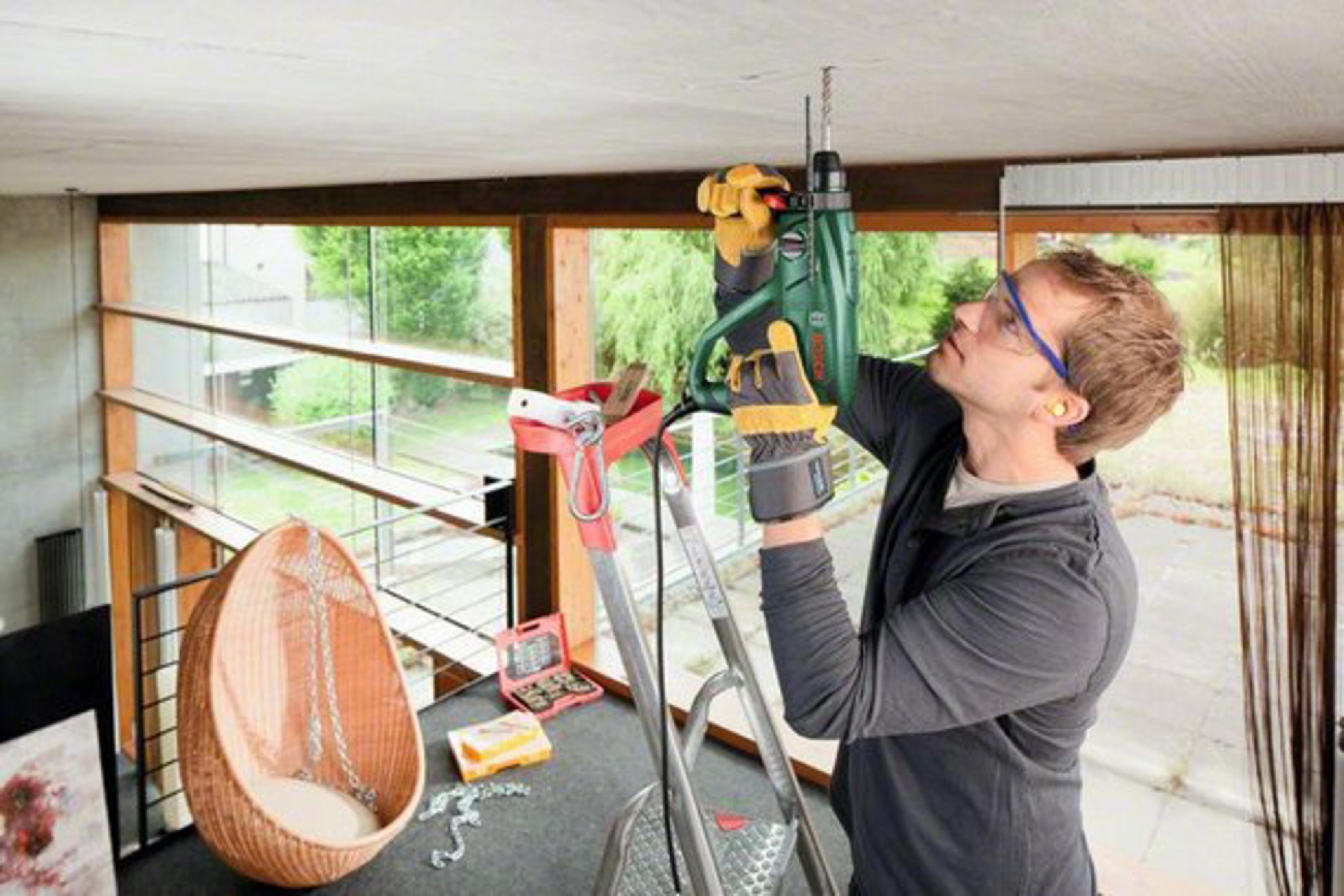 Ein Mann bohrt mit einem Bohrhammer in die Decke. Er trägt eine Schutzbrille und Ohrstöpsel und steht auf einer Leiter.