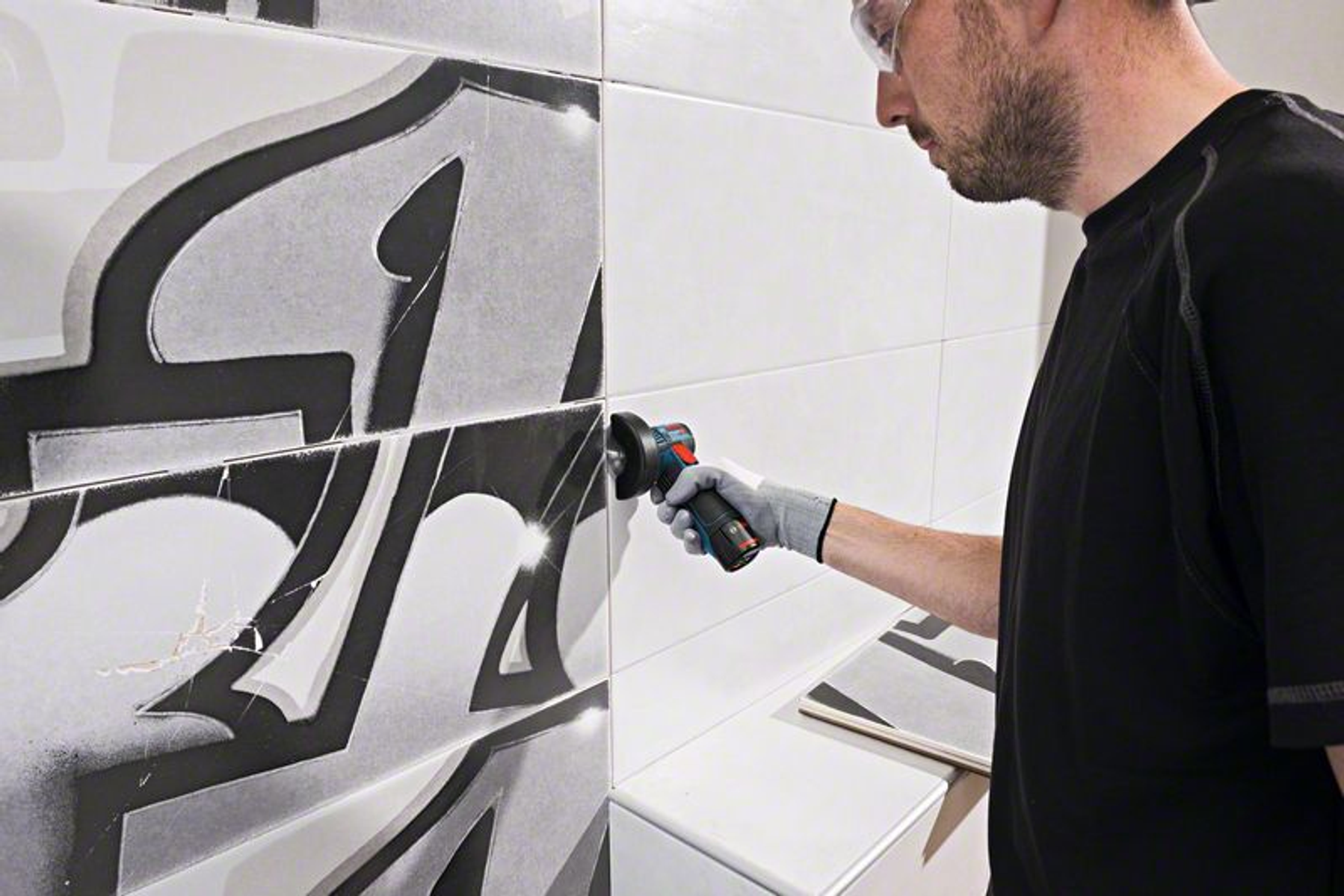 Mężczyzna używa szlifierki na kafelkowej ścianie. Płytki mają czarno-biały wzór. Mężczyzna ma okulary ochronne i rękawicę.