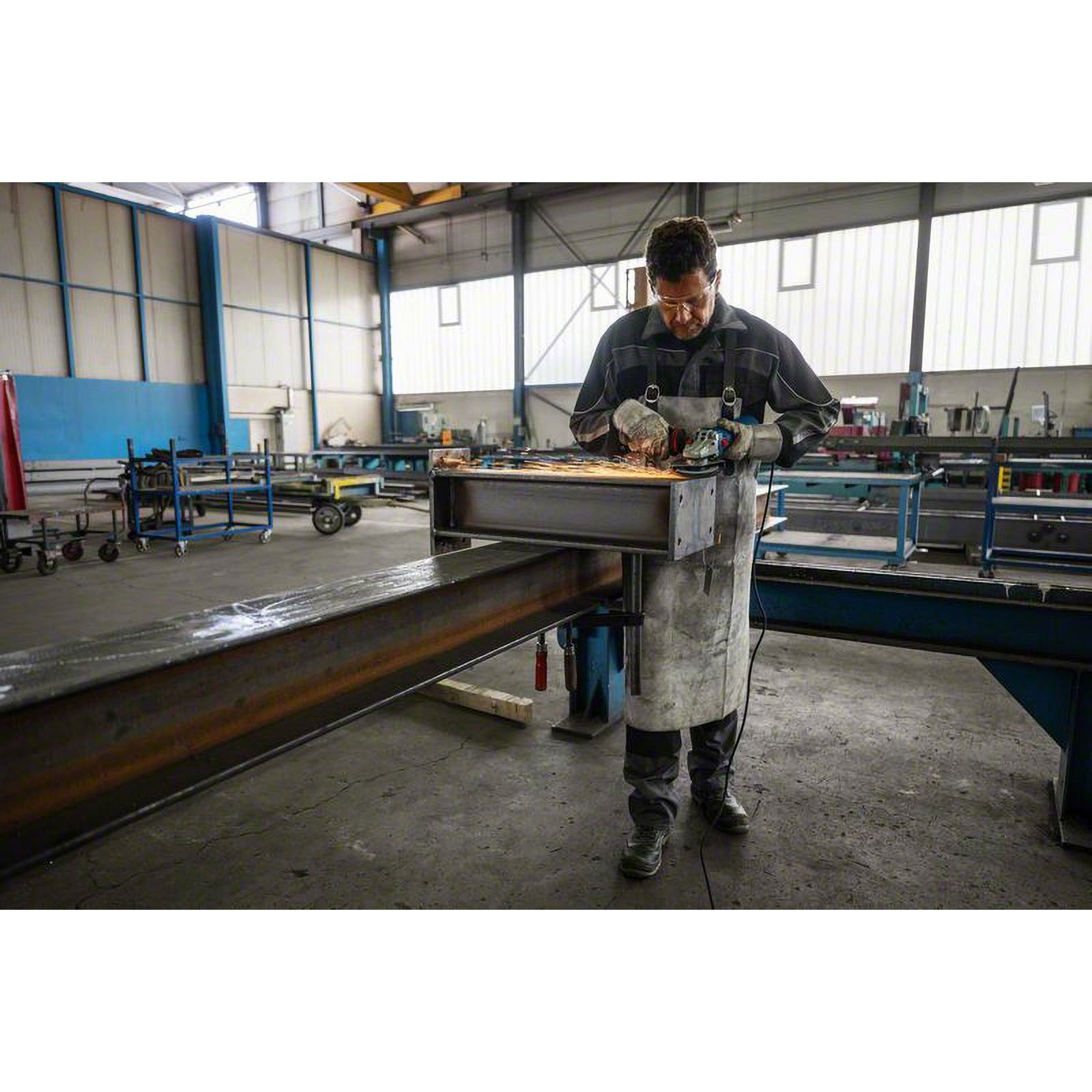 Bir adam atölyede metal bir kirişi taşlıyor. Kıvılcımlar uçuşuyor. Gözlük, eldiven, önlük ve tulum giyiyor.
