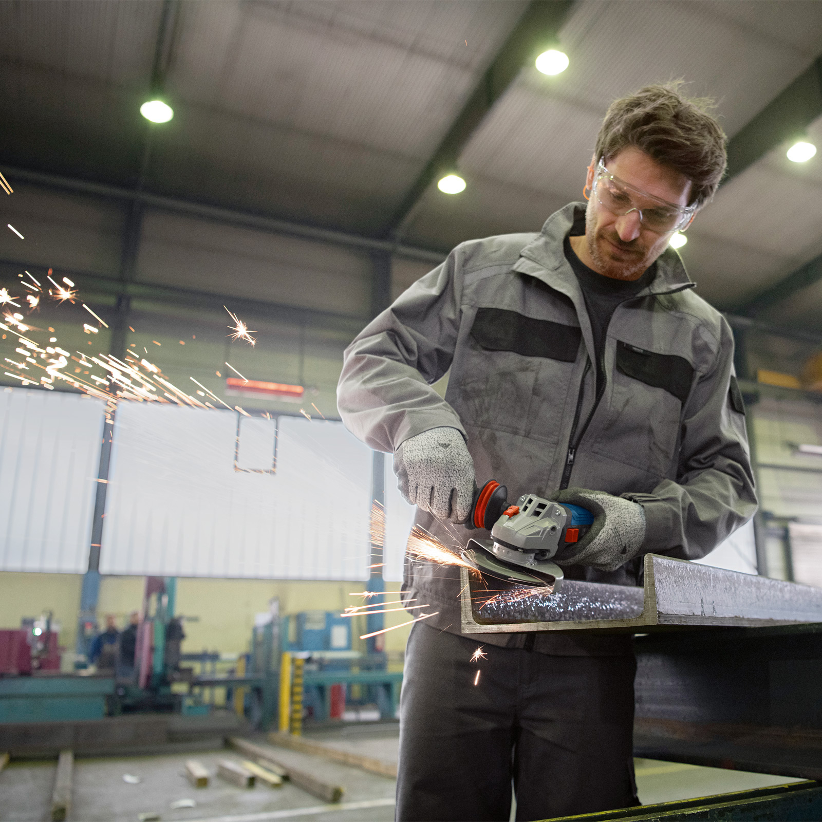 A man in gray workwear uses a grinder on metal in a workshop. Sparks fly. Safety glasses are worn.