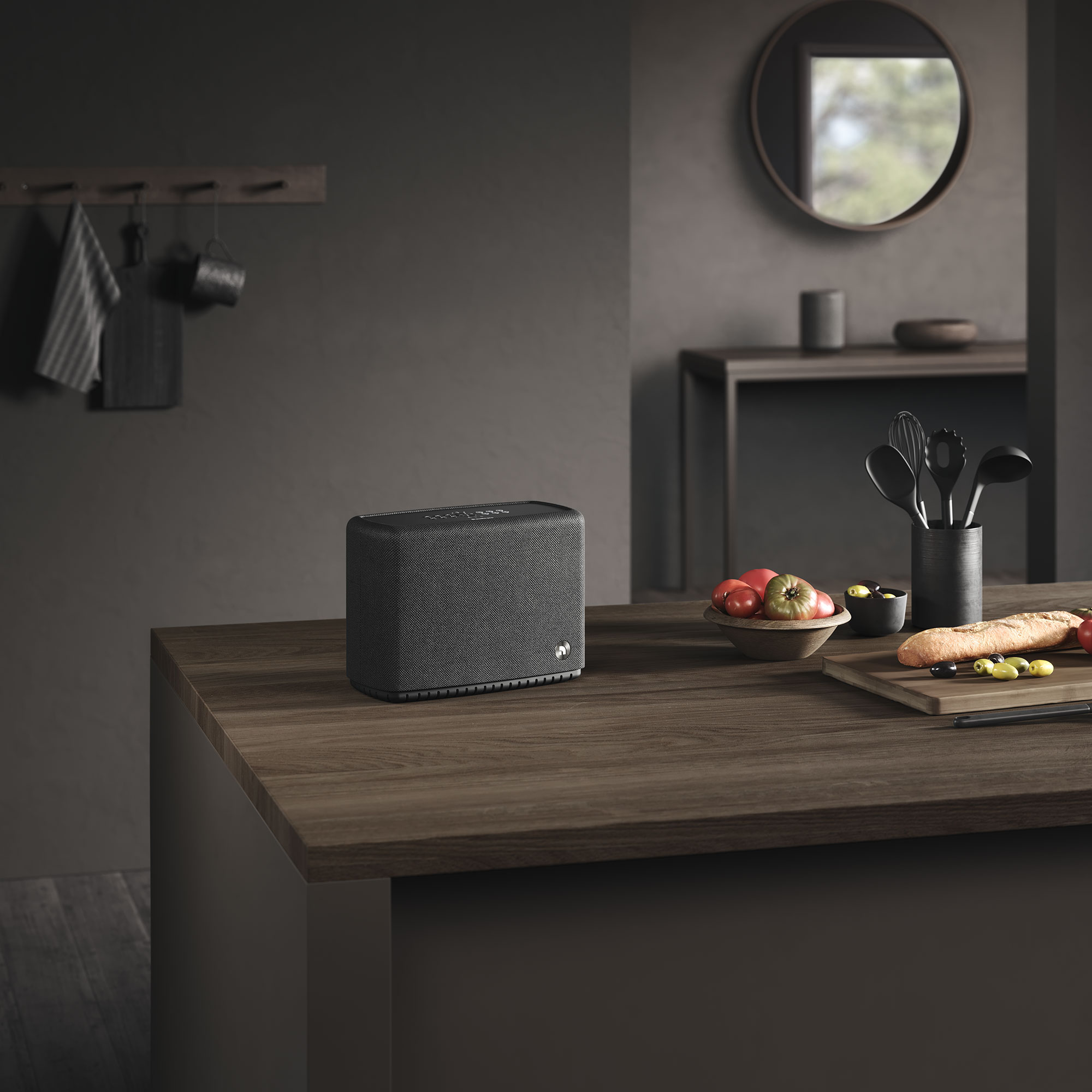 Ein Lautsprecher steht auf einer Holztheke mit Essen und Utensilien. Ein Spiegel und ein Ständer mit Gegenständen sind im Hintergrund.