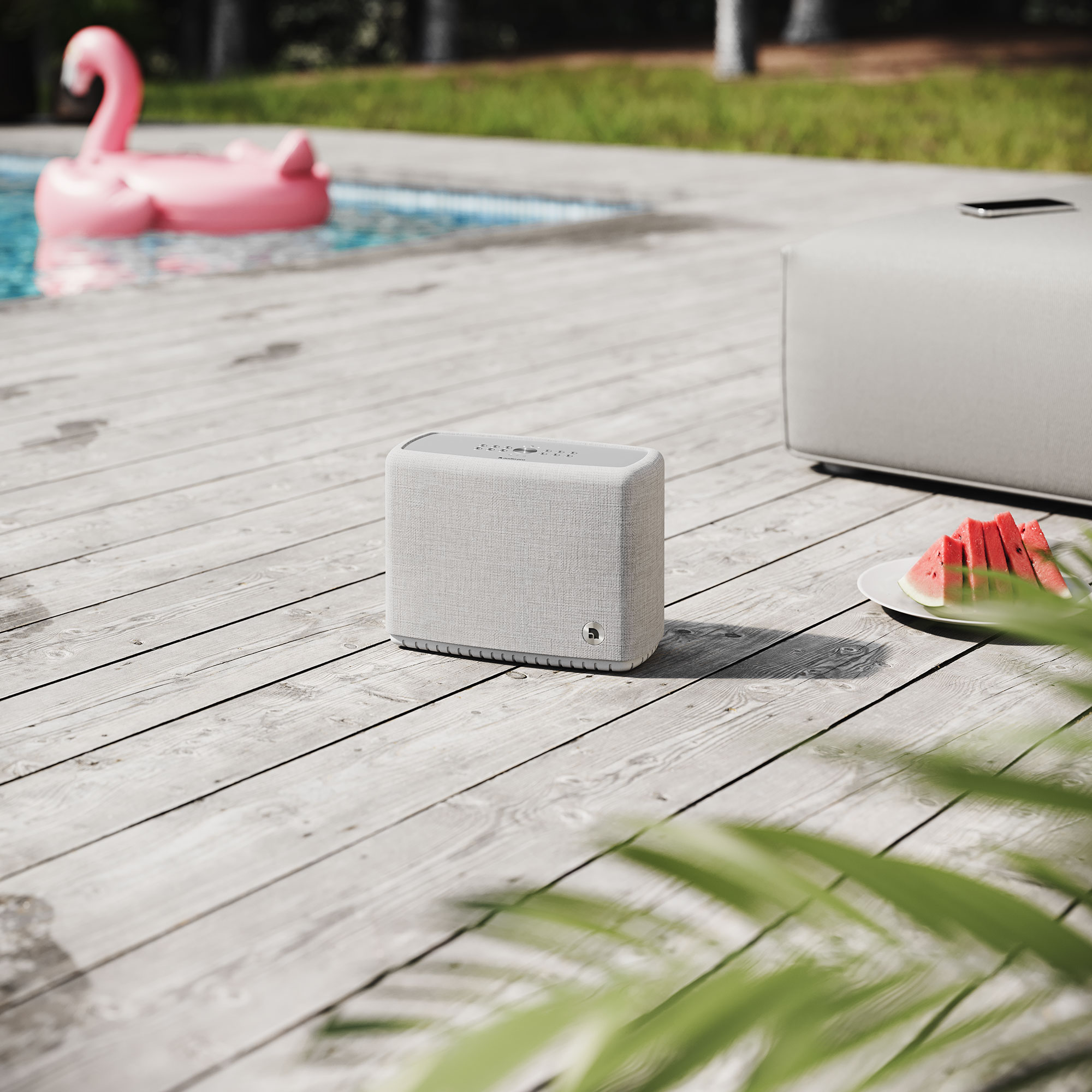 Un haut-parleur sur une terrasse en bois au bord d'une piscine. Un flotteur flamant rose. Des tranches de pastèque sur une assiette. Un téléphone sur un pouf blanc.