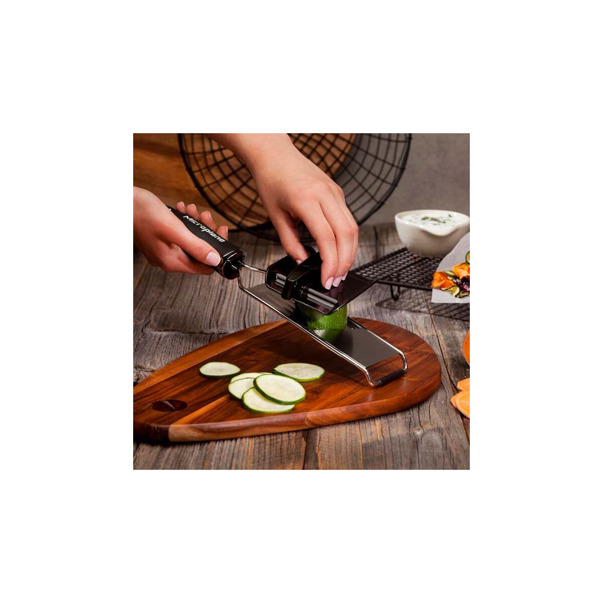 Eine Person raspelt Zucchini auf einem Schneidebrett. Geschnittene Zucchini und eine Schüssel mit Dip sind sichtbar. Ein schwarzer Reibeisen wird verwendet.