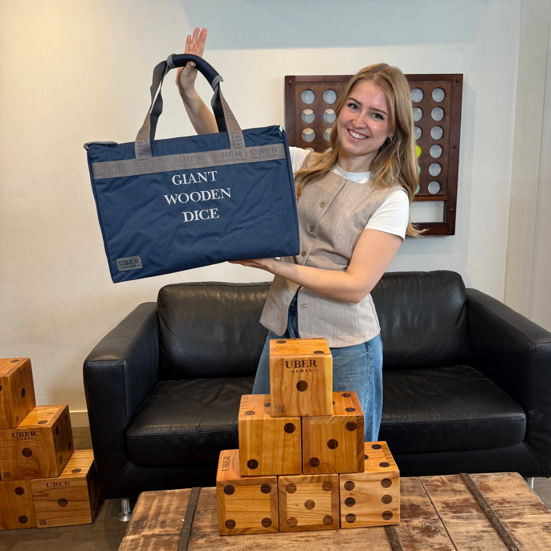 Eine Frau hält eine blaue Tasche mit der Aufschrift 'Giant Wooden Dice'. Im Hintergrund befinden sich Würfel und ein schwarzes Sofa.