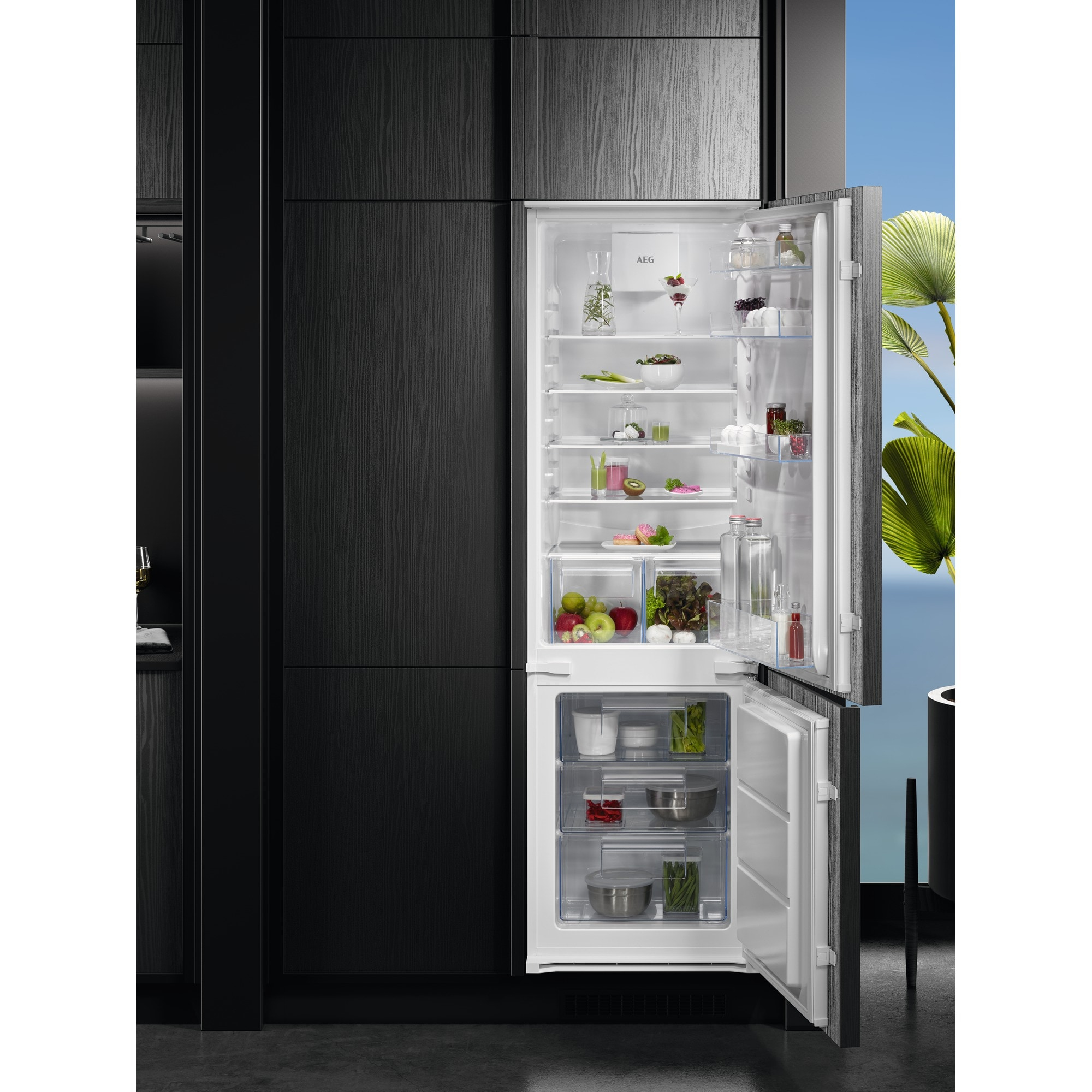 Un frigorífico moderno con las puertas abiertas, lleno de comida. Gabinetes negros enmarcan el refrigerador. Cielo azul brillante y vegetación al fondo.
