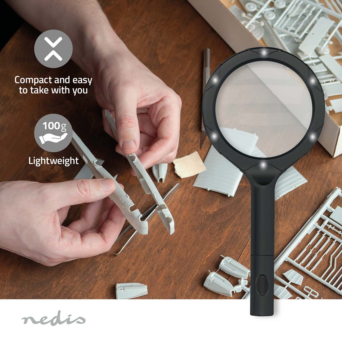Handen die een model in elkaar zetten met gereedschap en een vergrootglas op een houten oppervlak. Modelonderdelen en een doos op de achtergrond.