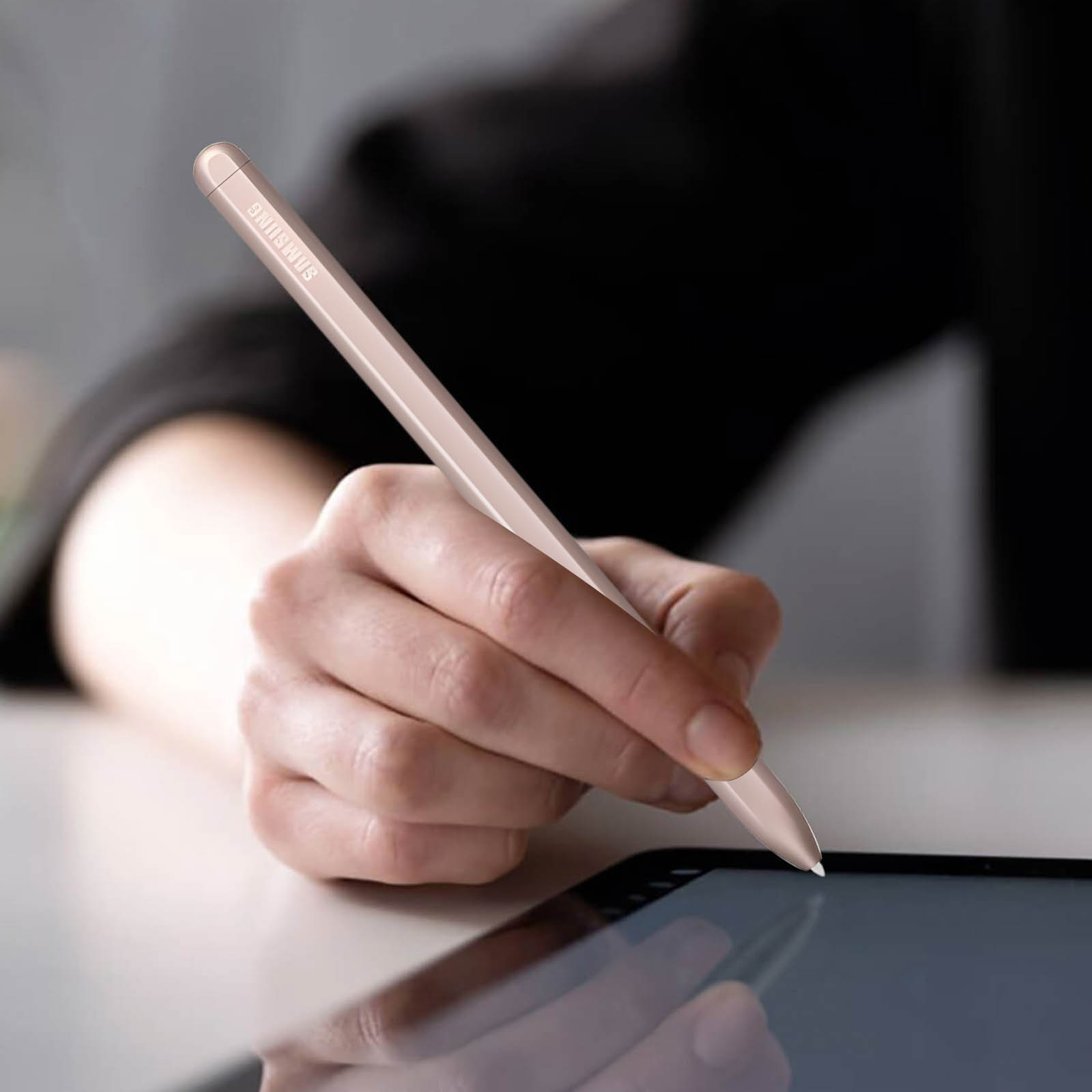 Eine Hand hält einen roségoldenen Samsung-Stift und schreibt auf ein Tablet. Der Hintergrund hat eine verschwommene Person in Schwarz.