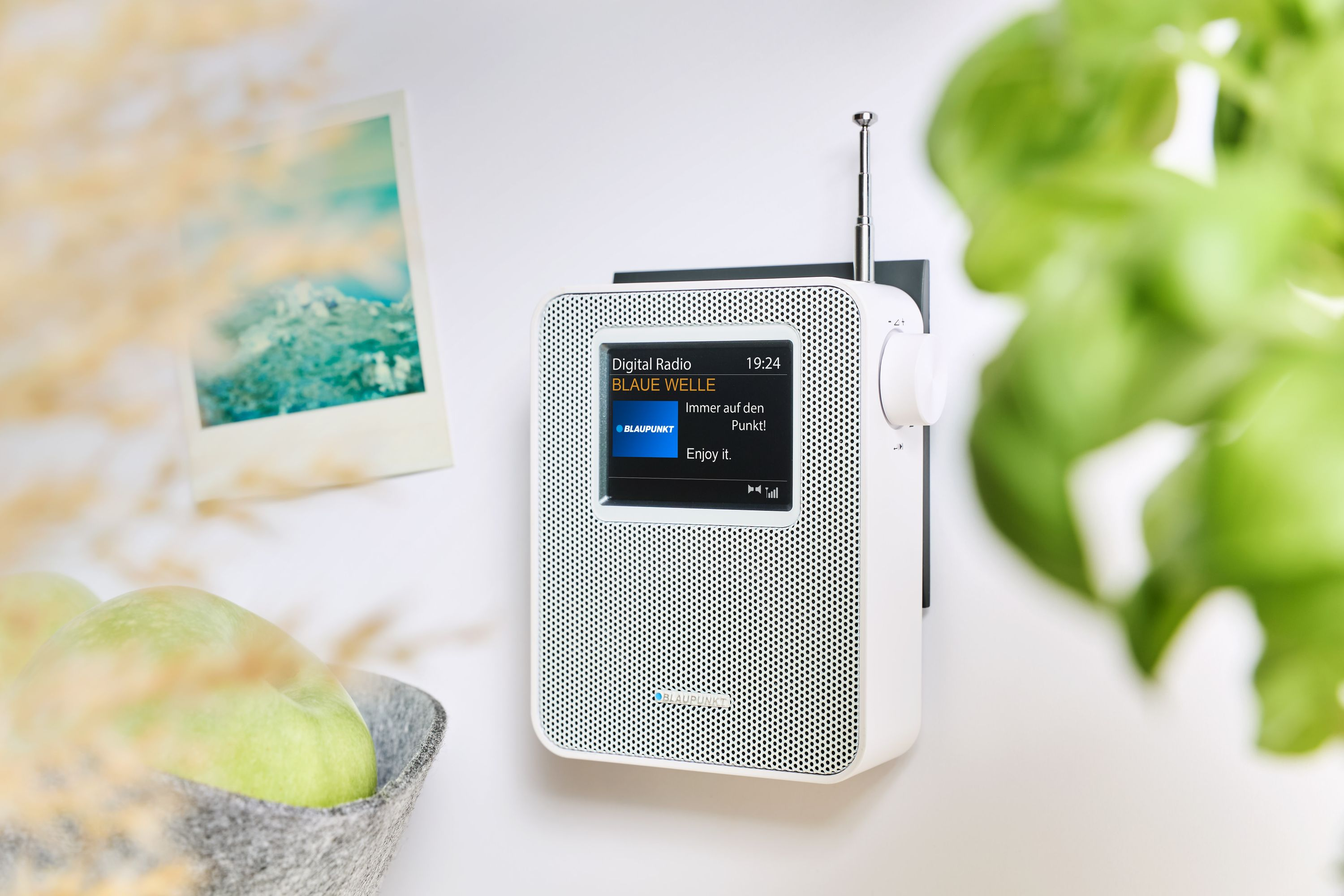 Ein weißes Digitalradio mit Bildschirm zeigt Blaupunkt. Daneben ein Foto und grüne Äpfel in einem Korb.