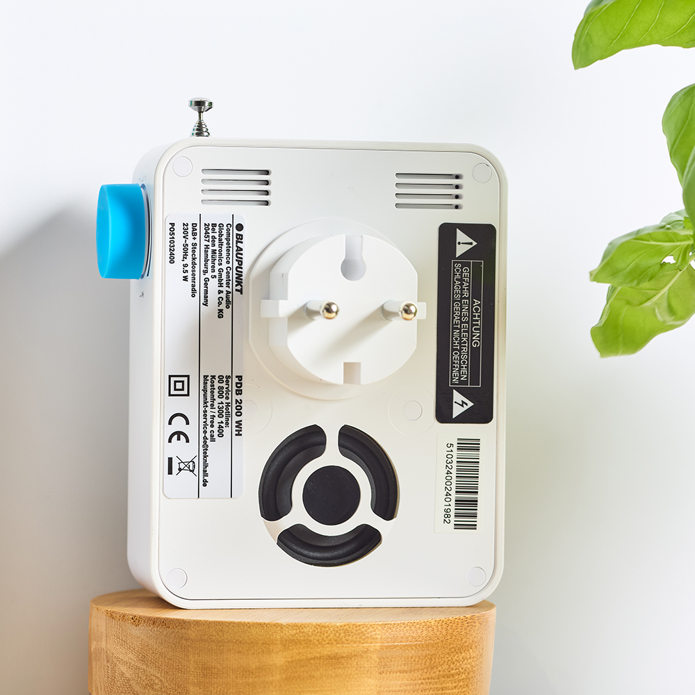 Ein weißes Blaupunkt-Radio mit Stecker, blauem Anschluss und Lautsprecher auf einem Holzständer. Grüne Blätter im Hintergrund.