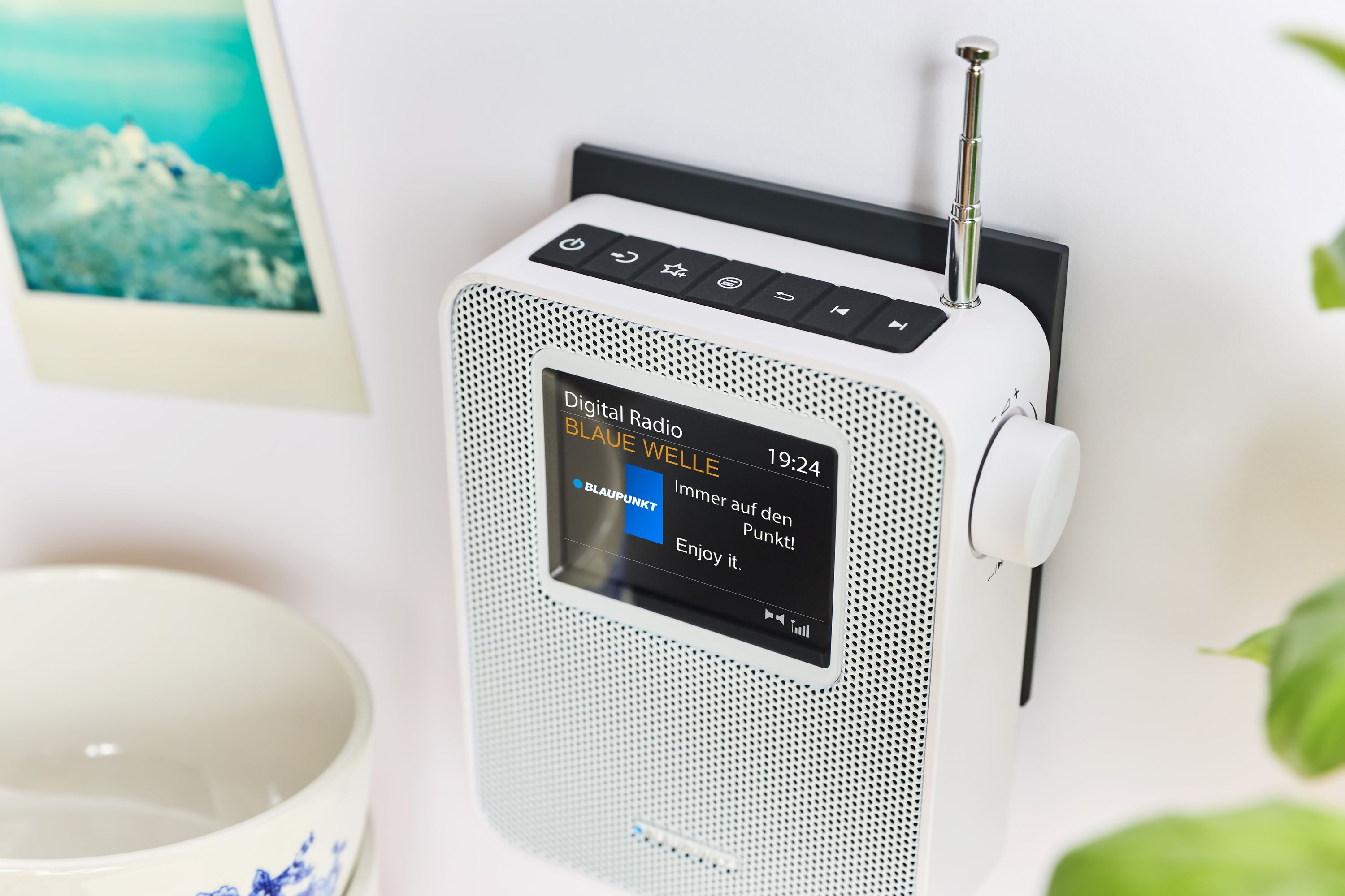 Ein weißes Digitalradio an der Wand. Es zeigt einen Radiosender an. Ein Polaroid-Foto und eine Schüssel sind im Bild.