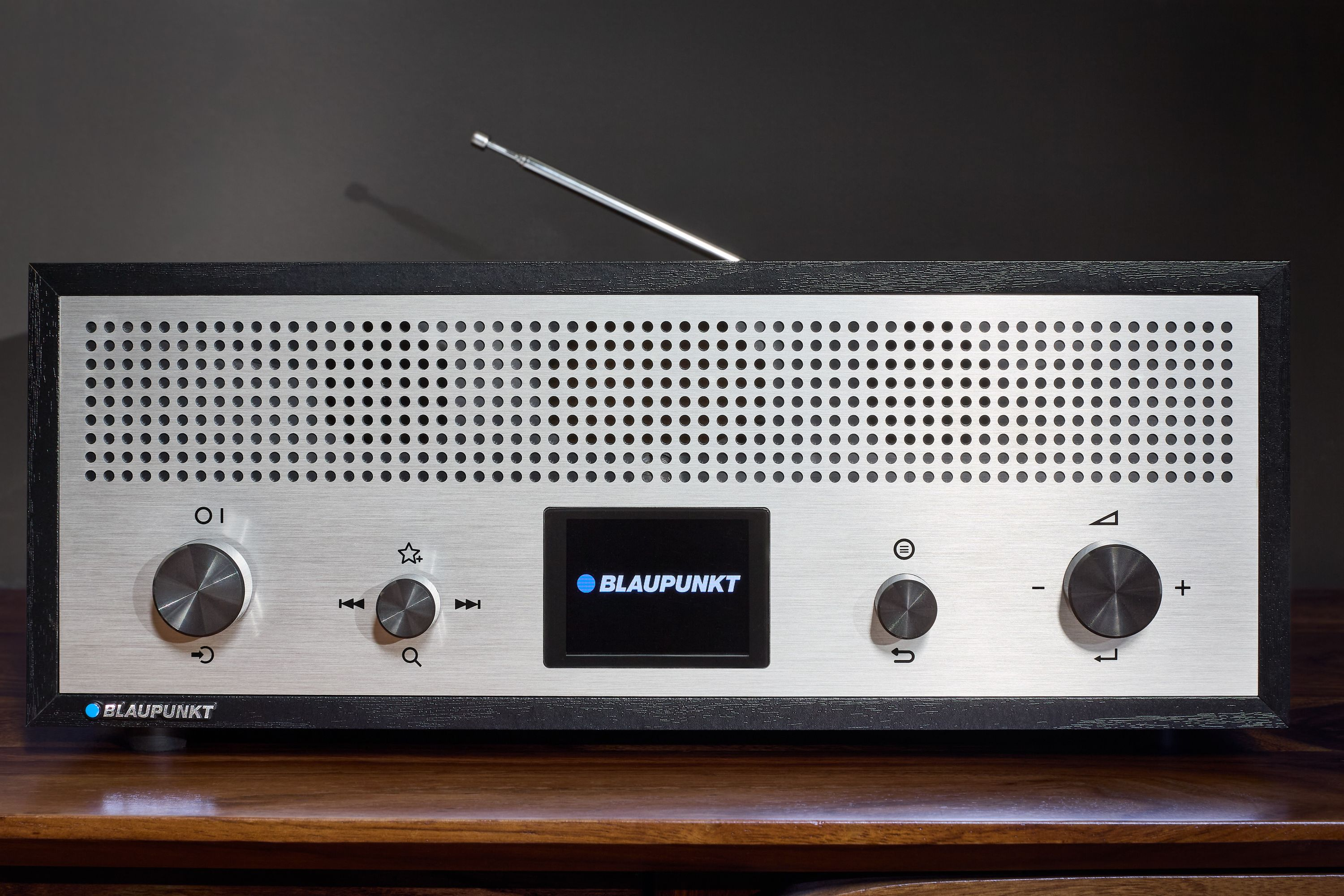 Silbernes Blaupunkt-Radio mit schwarzem Rahmen, Knöpfen, Bildschirm und Antenne auf einer Holzoberfläche vor dunklem Hintergrund.