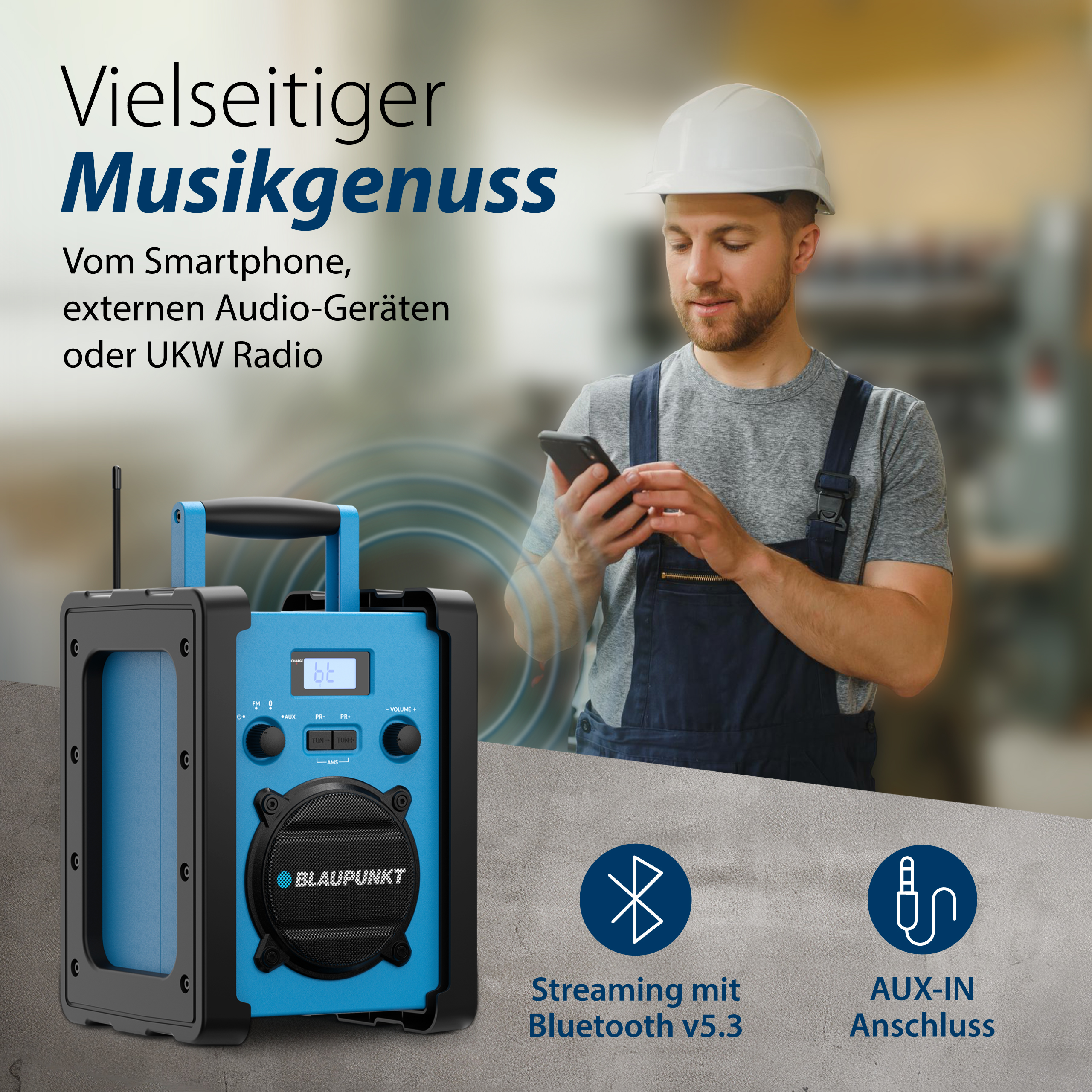 Ein Mann in Overalls und Helm benutzt ein Telefon. Ein blau-schwarzer Lautsprecher ist im Vordergrund.