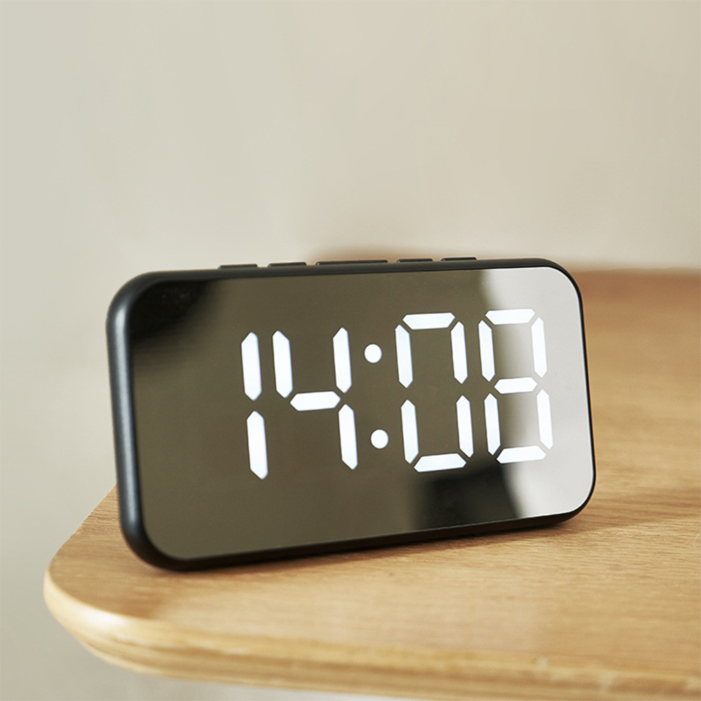 Eine Digitaluhr zeigt 14:08 auf einer reflektierenden schwarzen Oberfläche. Sie liegt auf einem Holzregal vor neutralem Hintergrund.