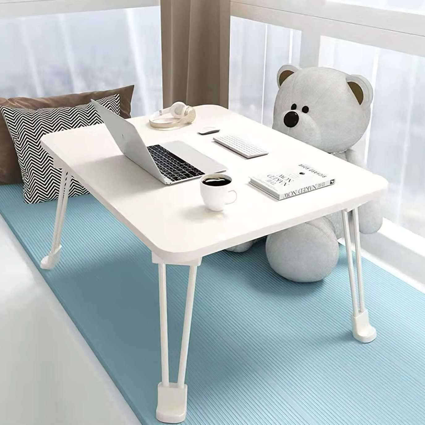 Ein Laptop steht auf einem weißen Klapptisch. Eine Kaffeetasse, ein Buch und ein Teddybär befinden sich auf dem Tisch. Kissen und ein Fenster sind im Hintergrund.