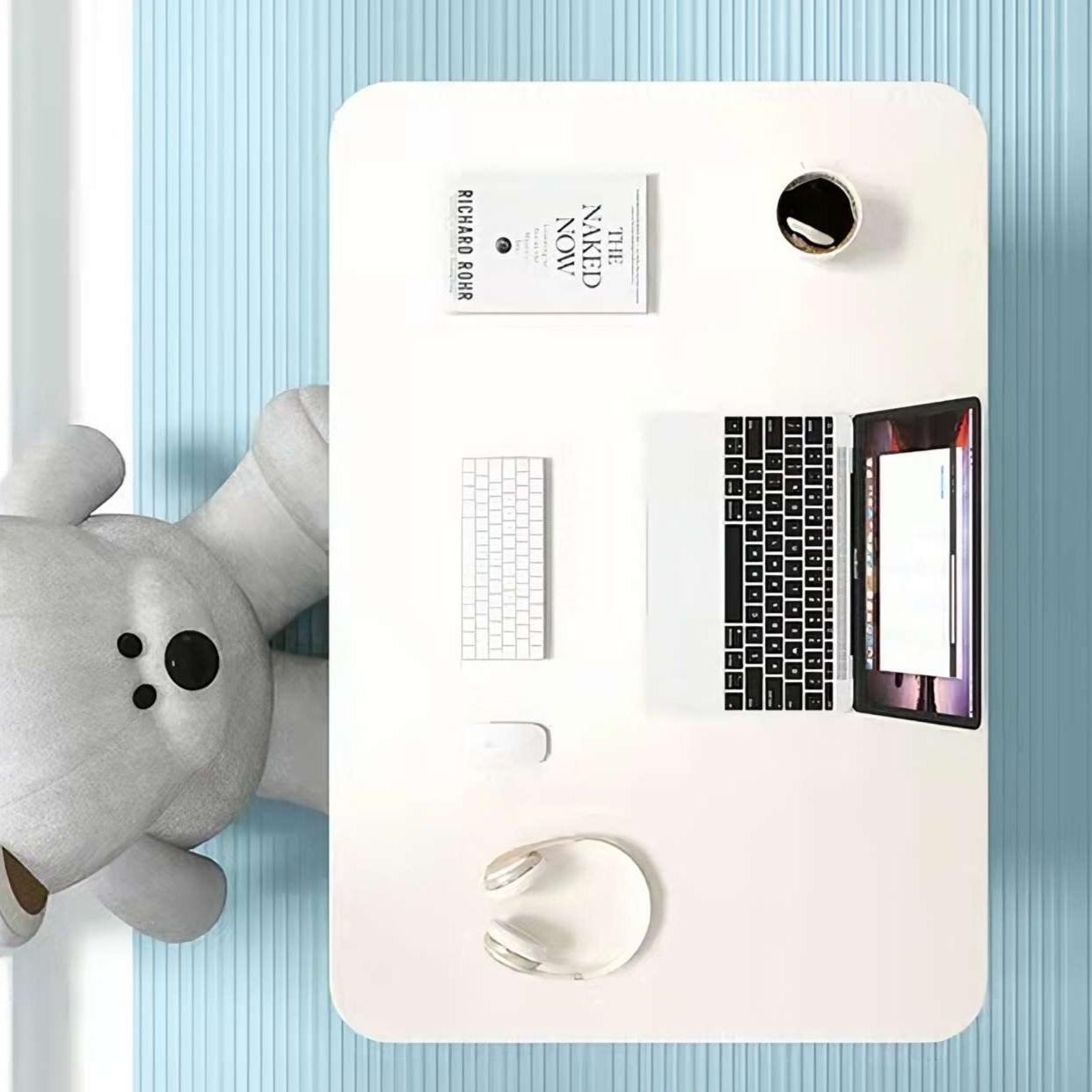 Weißer Schreibtisch mit Laptop, Tastatur, Kaffee, Buch und Kopfhörern. Ein Plüschbär ist links. Blauer, gestreifter Wandhintergrund.