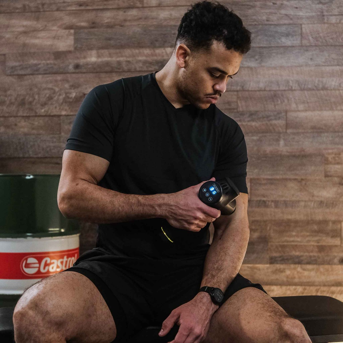 Mężczyzna w czerni używa pistoletu do masażu na ramieniu. Siedzi na ławce, z zielonym bębnem i drewnianymi panelami w tle.