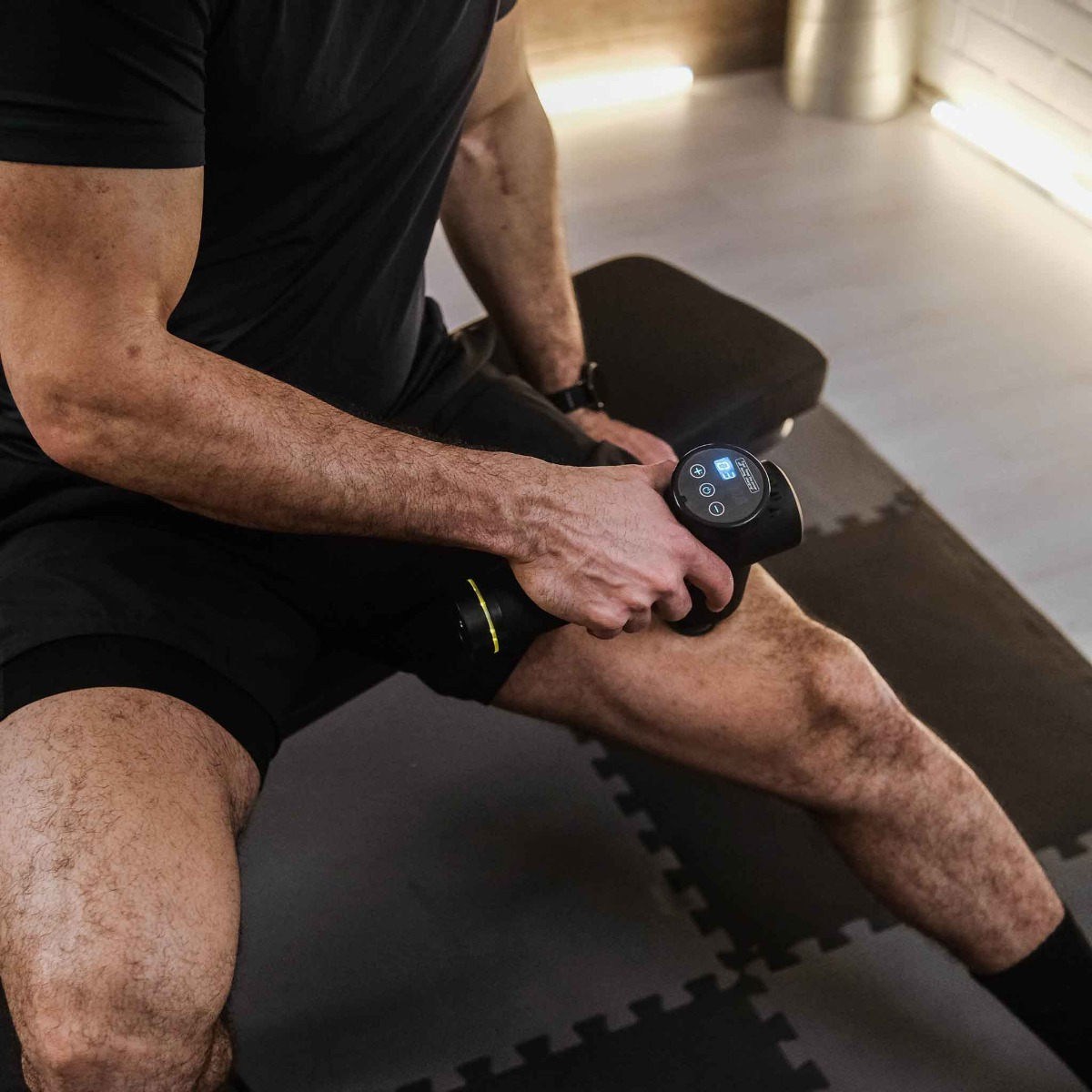 Mężczyzna w czarnych szortach używa pistoletu do masażu na udzie w pozycji siedzącej. Tło to sala gimnastyczna.