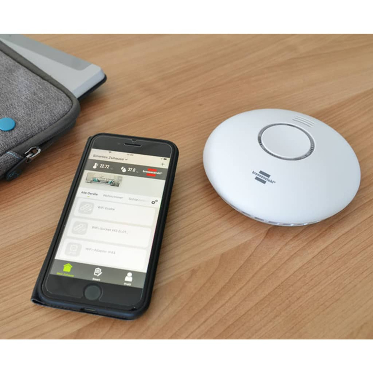 Un smartphone muestra una aplicación de hogar inteligente junto a un detector de humo blanco sobre una superficie de madera. Un estuche gris es parcialmente visible.
