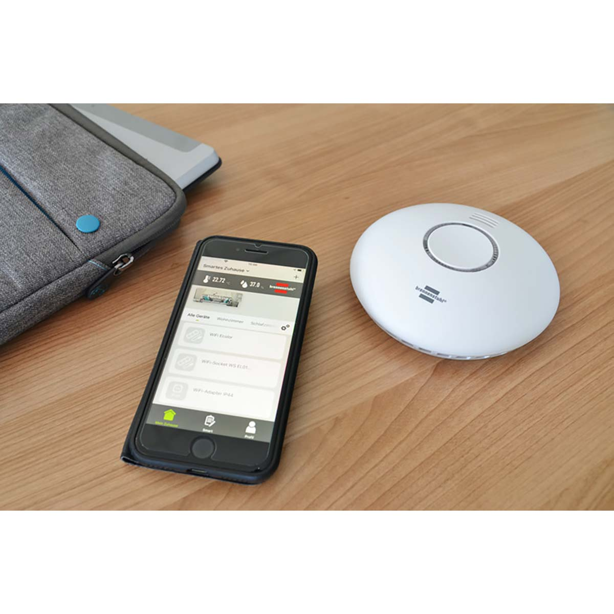 Un smartphone muestra controles de hogar inteligente junto a un dispositivo blanco y circular sobre una superficie de madera. En el fondo hay una funda gris para portátil.