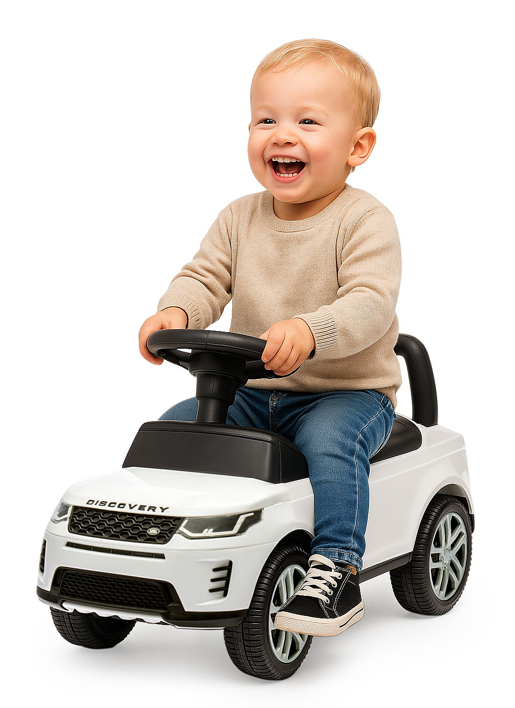 Un niño pequeño sonriente se sienta en un coche de juguete blanco, sujetando el volante. Lleva vaqueros y un suéter beige.