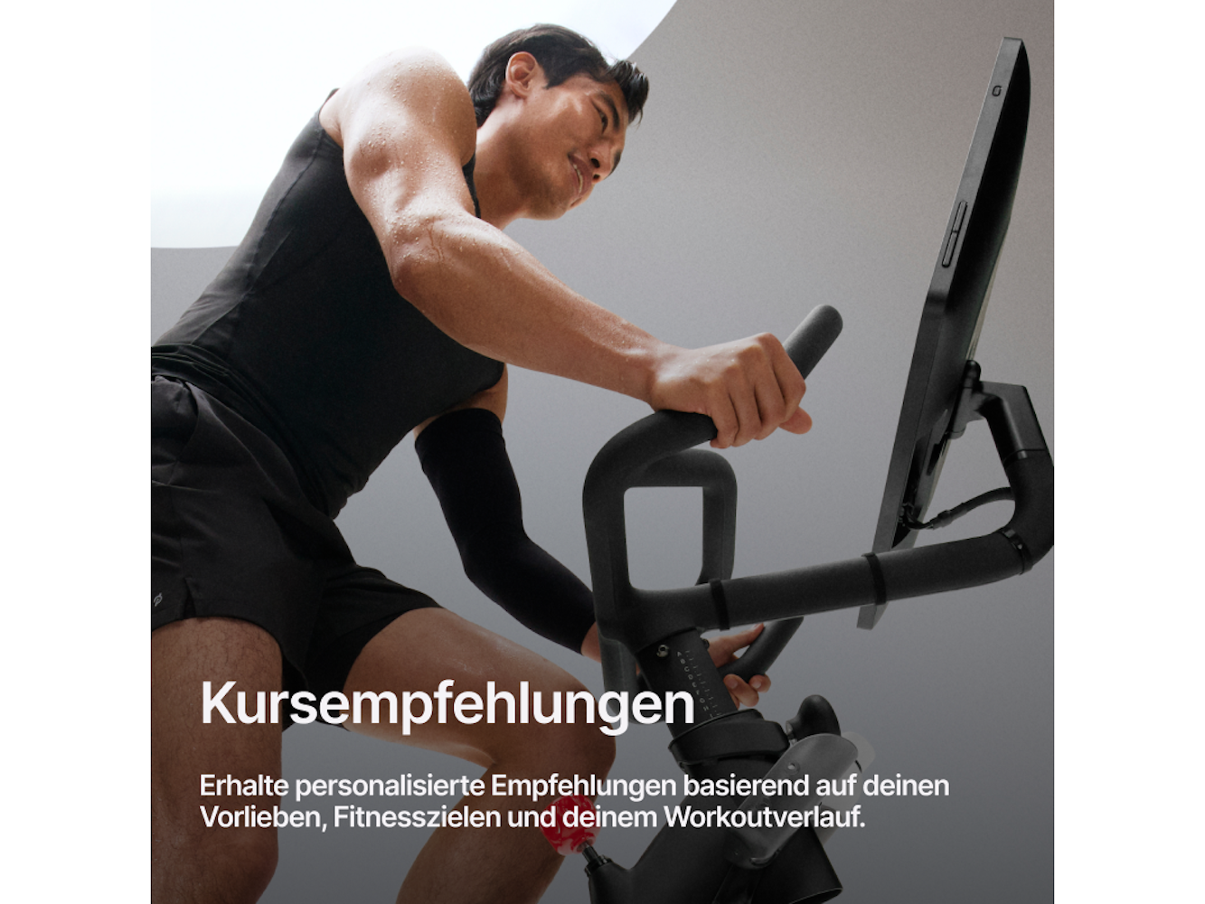 Ein Mann trainiert auf einem Heimtrainer. Er trägt schwarze Trainingskleidung. Das Fahrrad hat einen Bildschirm und Griffe.