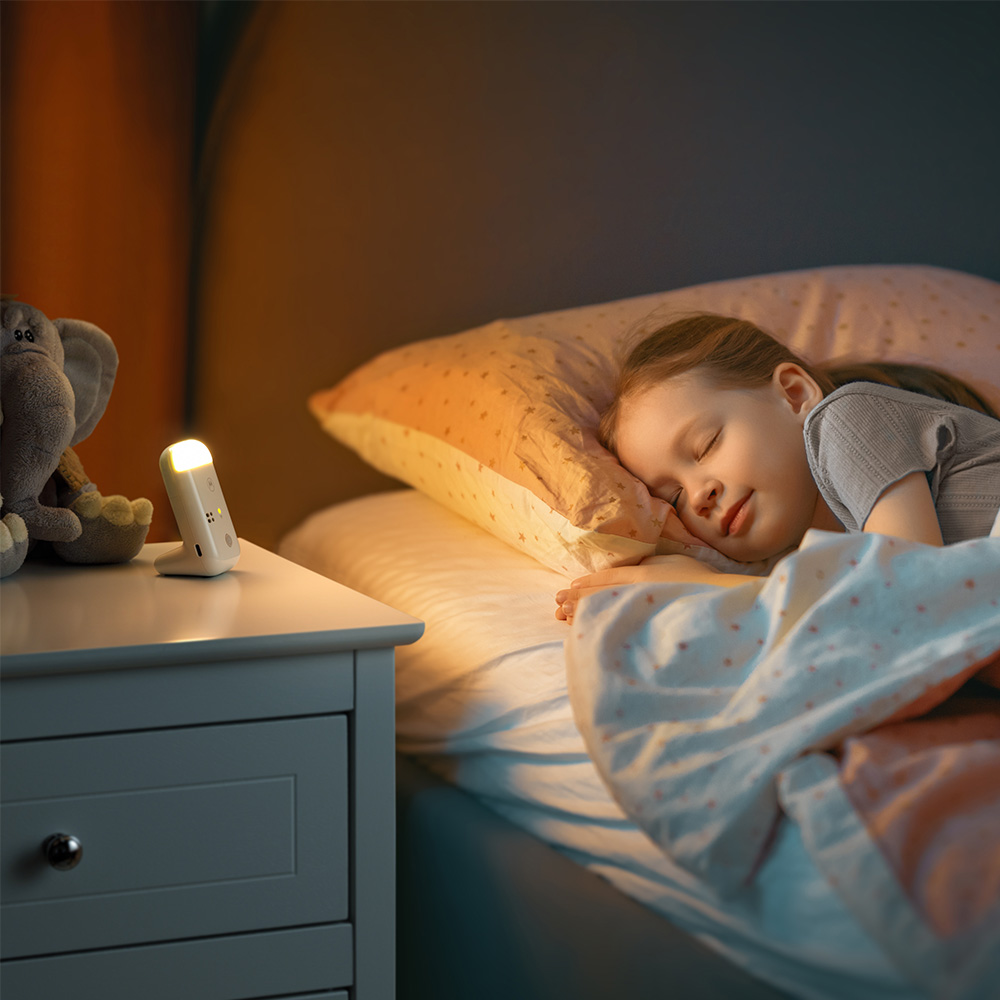 Una niña duerme en la cama. Una luz nocturna encendida y un elefante de peluche están en una mesita de noche. La escena está tenue.