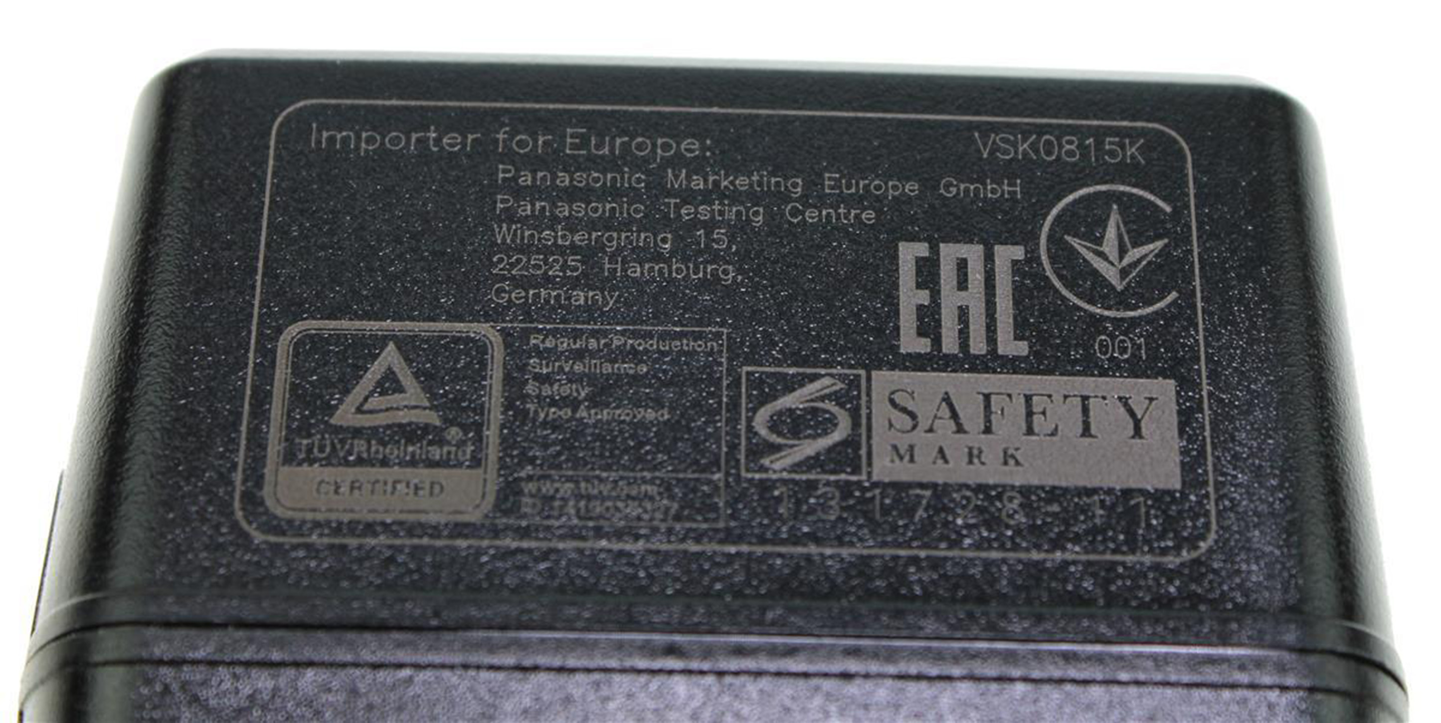 Detail eines schwarzen Netzteils. Text enthält Importeurinformationen, Sicherheitszeichen und Zertifizierungen. Grauer Text auf schwarzem Grund.