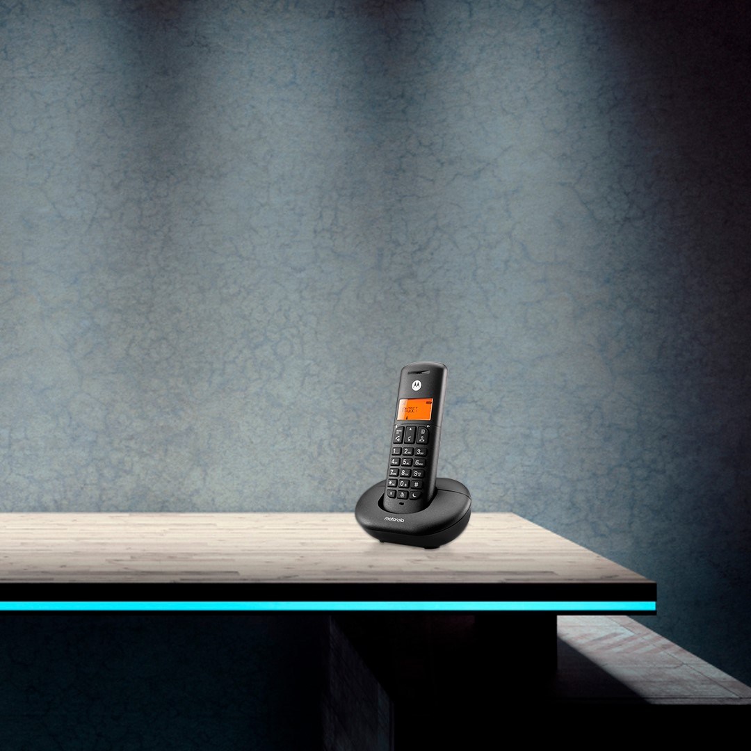 Un teléfono inalámbrico Motorola negro descansa en su base de carga sobre una superficie de madera clara contra una pared gris texturizada.