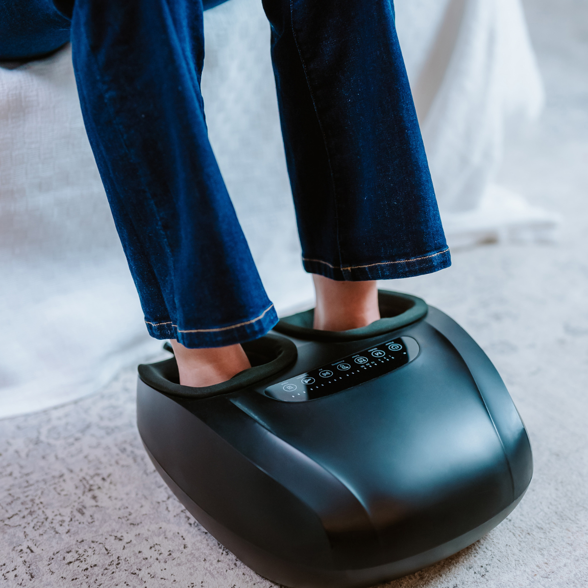 Füße einer Person in einem schwarzen Fußmassagegerät. Blaue Jeans. Das Massagegerät hat Knöpfe und einen Bildschirm. Auf hellem Teppich.