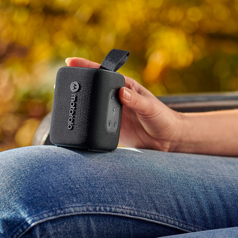 Eine Person hält einen schwarzen Motorola-Lautsprecher auf dem Schoß. Der Hintergrund zeigt Herbstlaub. Die Person trägt Jeans.