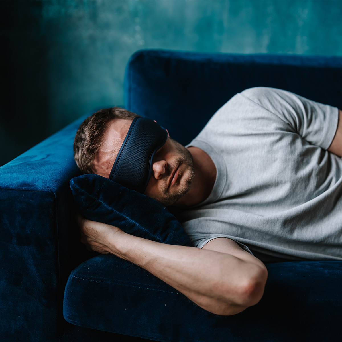 Ein Mann mit Schlafmaske liegt auf einem blauen Sofa und stützt seinen Kopf auf ein Kissen. Er trägt ein graues Hemd.