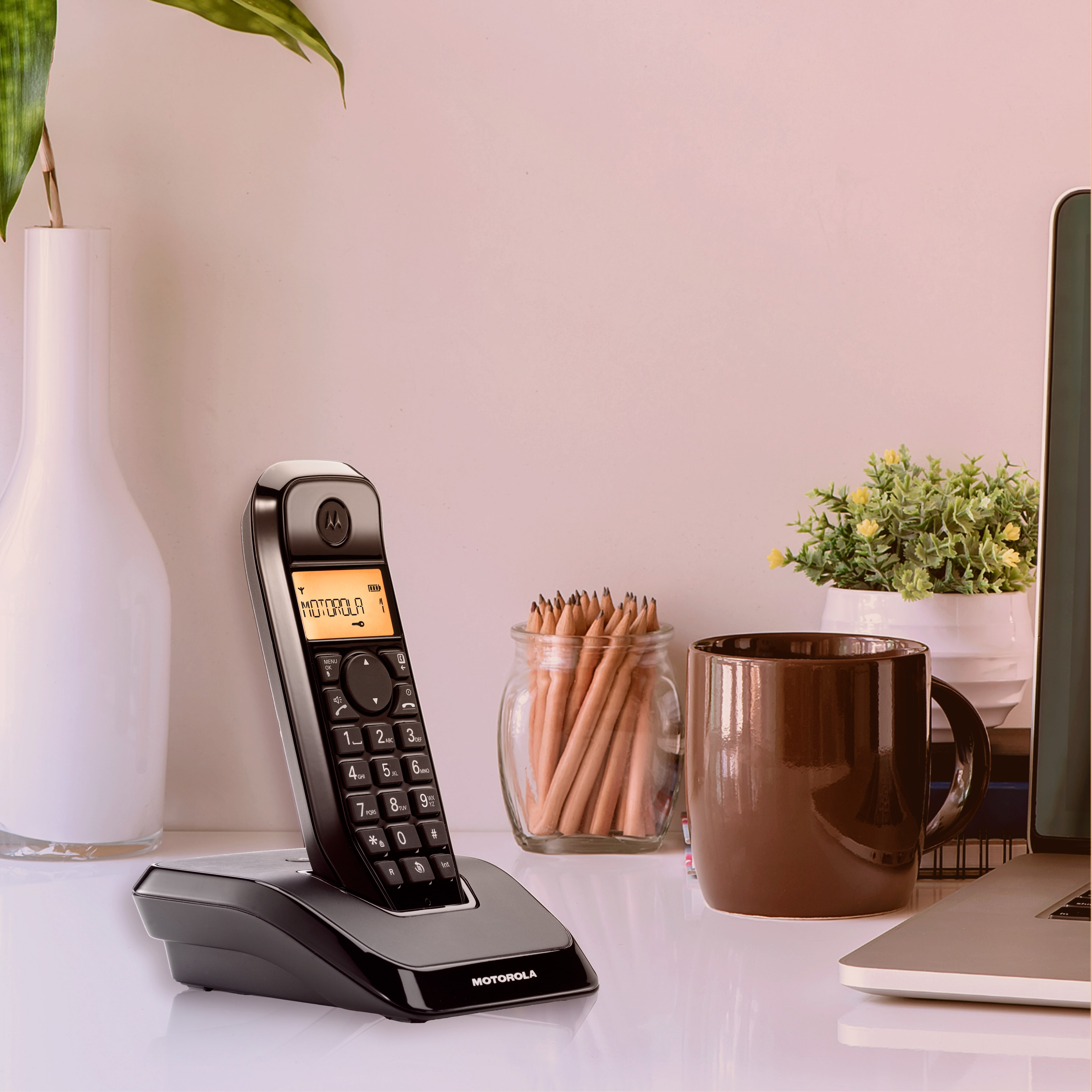 Ein schwarzes Motorola-Schnurlostelefon auf einem weißen Schreibtisch neben einer Kaffeetasse, Bleistiften und einem Laptop.