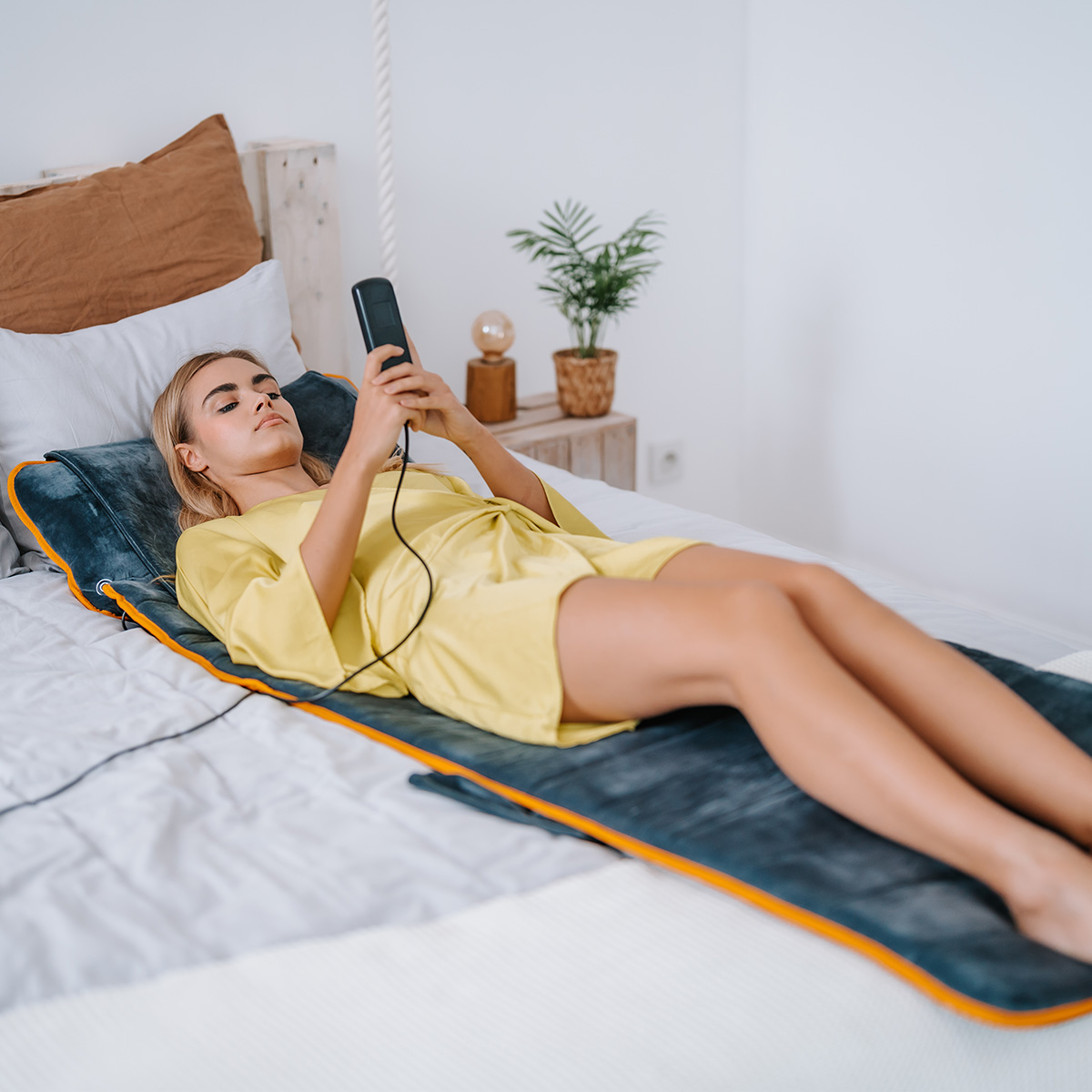 Mujer con bata amarilla acostada en la cama, usando un dispositivo. Cama con alfombra gris con borde naranja. Fondo: planta, lámpara y pared blanca.