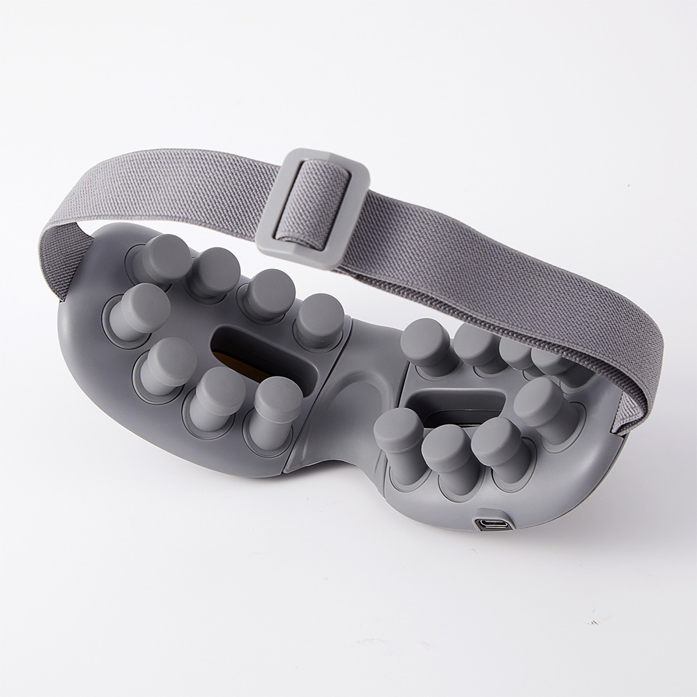 Un masajeador ocular gris con nodos salientes y una correa ajustable. El dispositivo tiene un puerto USB.