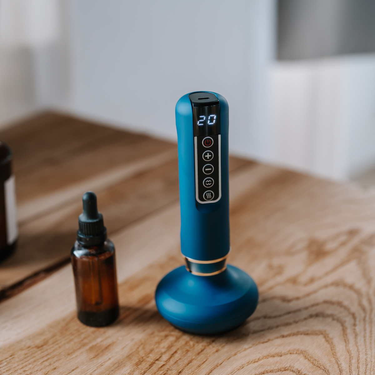 Un dispositivo de masaje azul con pantalla digital '20' y botones de control, junto a una botella marrón sobre una superficie de madera.