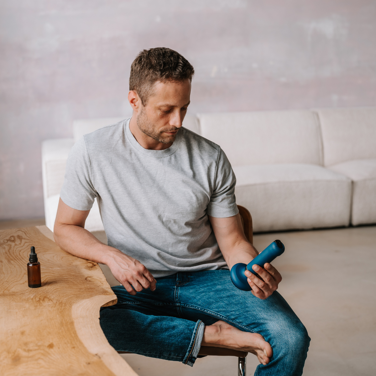 Un hombre con camisa gris y jeans sentado, mirando un objeto azul en su mano. Hay una botella en una mesa de madera.