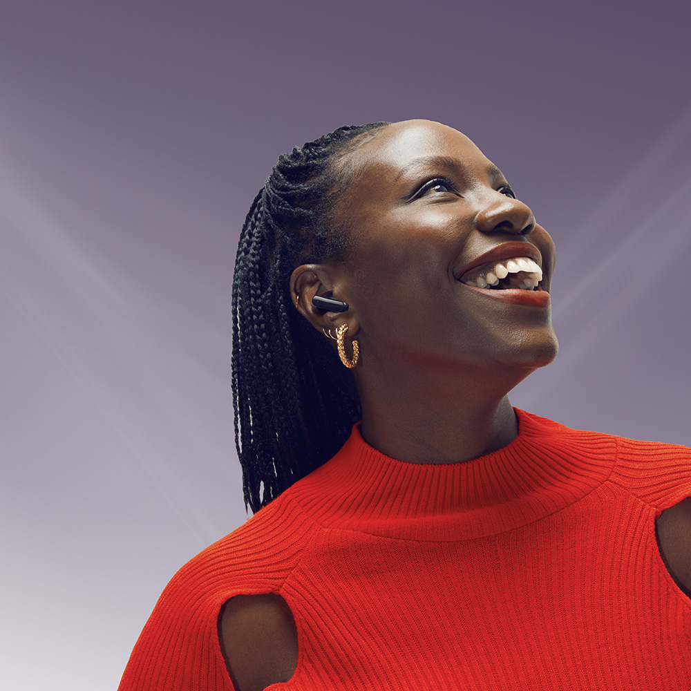 Una mujer sonriente con un suéter rojo, aretes dorados y un auricular negro, mirando hacia arriba. El fondo es un degradado púrpura.